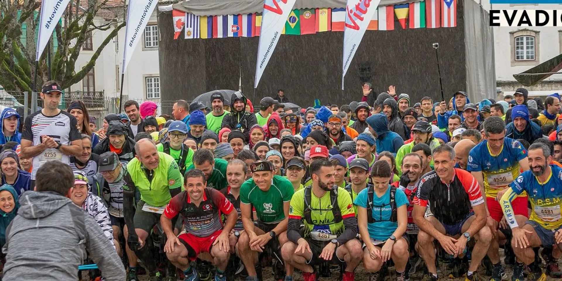 The image shows a large group of people gathered, many in athletic gear, likely participating in a race or sporting event. It's raining, and there's a stage in the background with flags of various countries. The participants are posing for a photo under several branding banners.