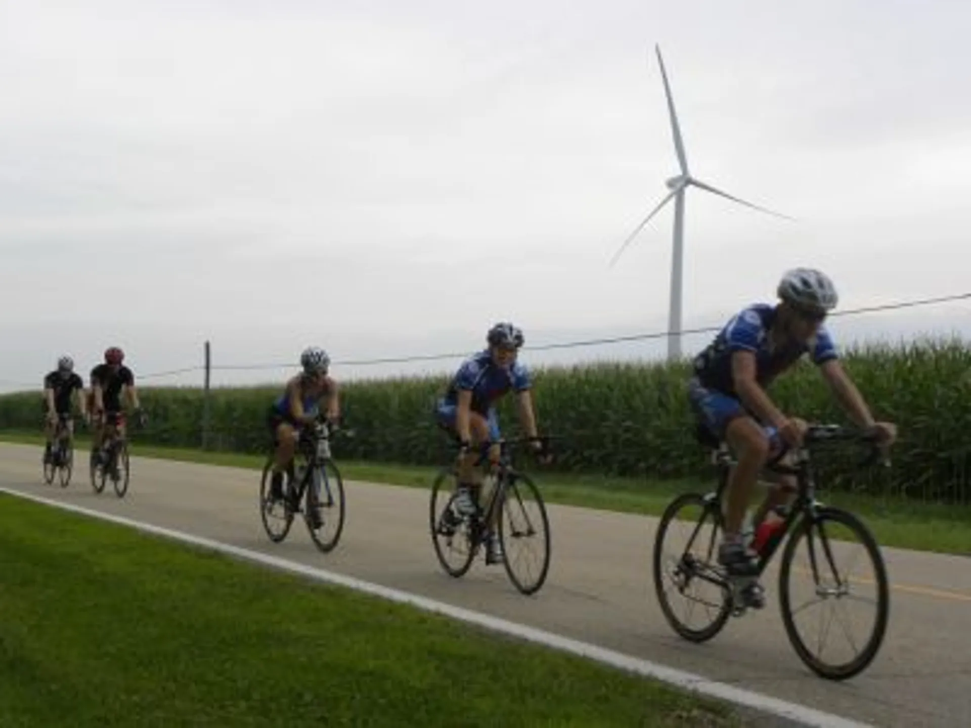 The image shows a group of cyclists riding on a road next to a field. In the background, there is a large wind turbine, indicating the presence of a wind farm or a rural area where renewable energy is harvested. The landscape appears to be quite flat, and the sky is overcast, suggesting an overcast day. The cyclists are wearing helmets and athletic clothing suitable for cycling.