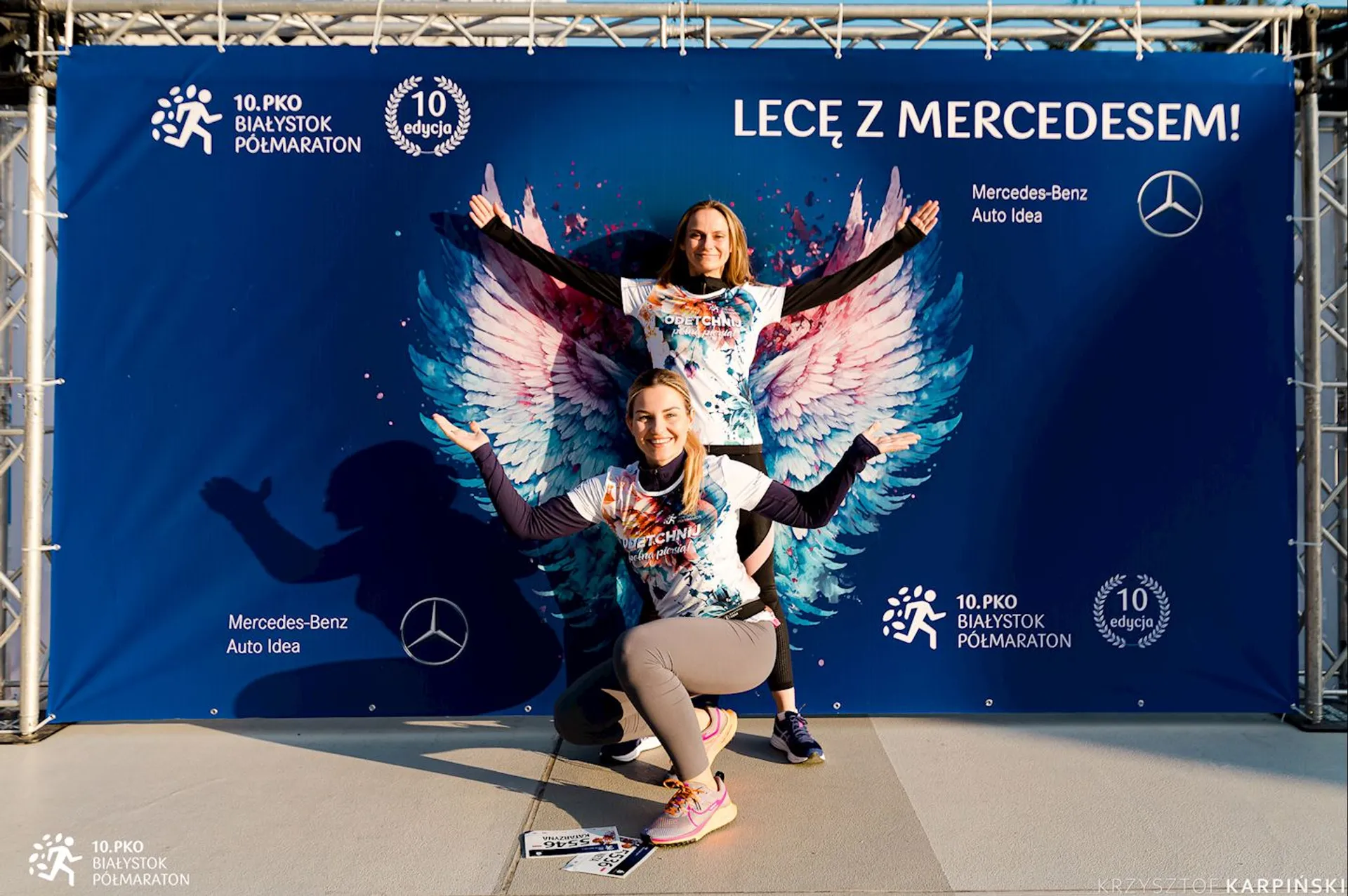 In this image, there are two people posing in front of a backdrop with a