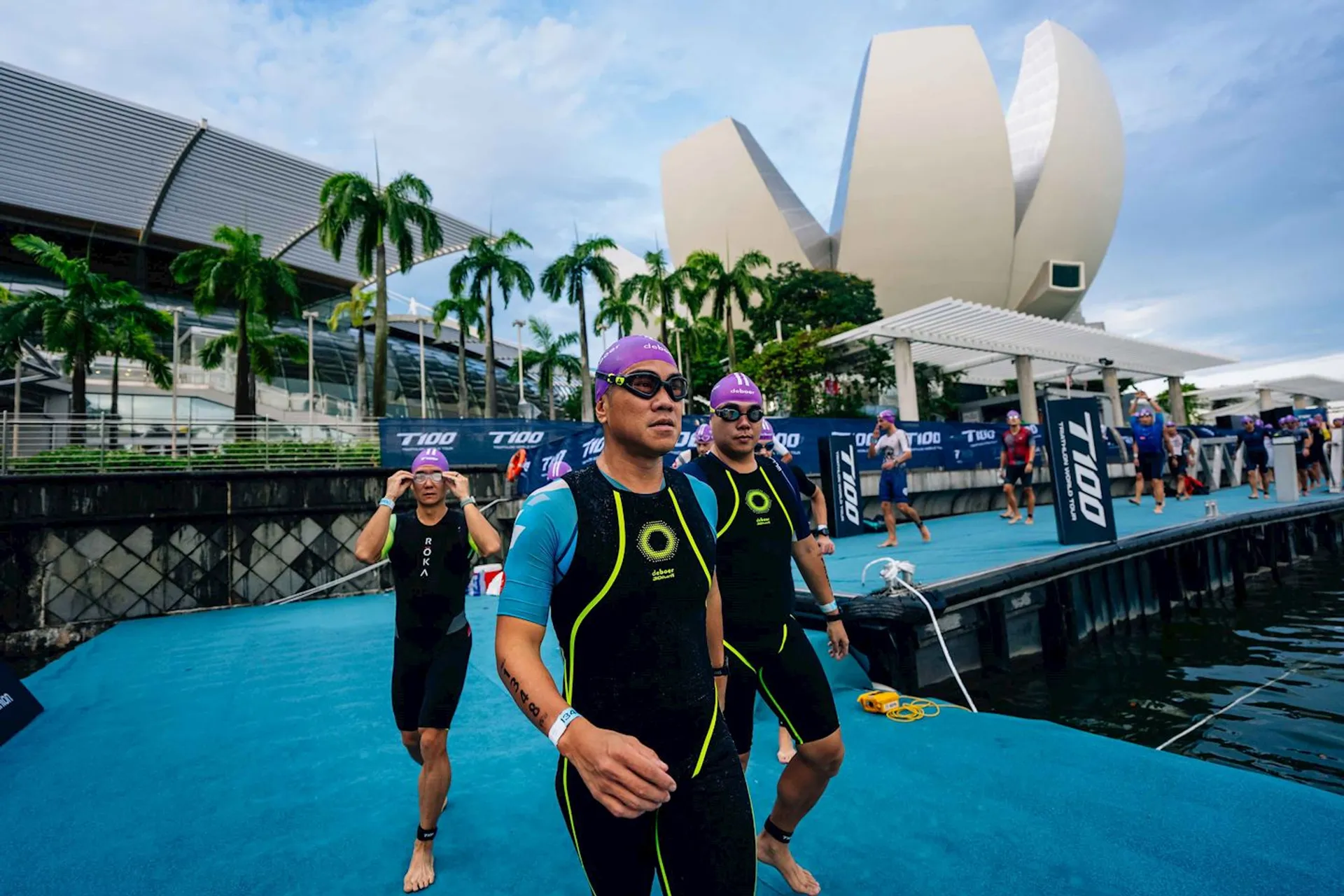 The image shows a group of people wearing swim caps and wetsuits, preparing for or finishing a swimming event. They are on a dock or platform near a body of water. In the background, there are modern buildings with distinctive architecture, palm trees, and event signage. It looks like a triathlon or a similar sporting competition.