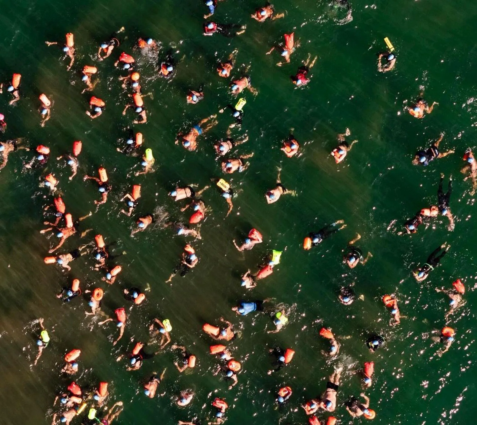 The image shows numerous people swimming in a body of water. Many are wearing orange and yellow flotation devices or life vests. The photo appears to be taken from above, capturing a lively scene with people spread across the water surface.