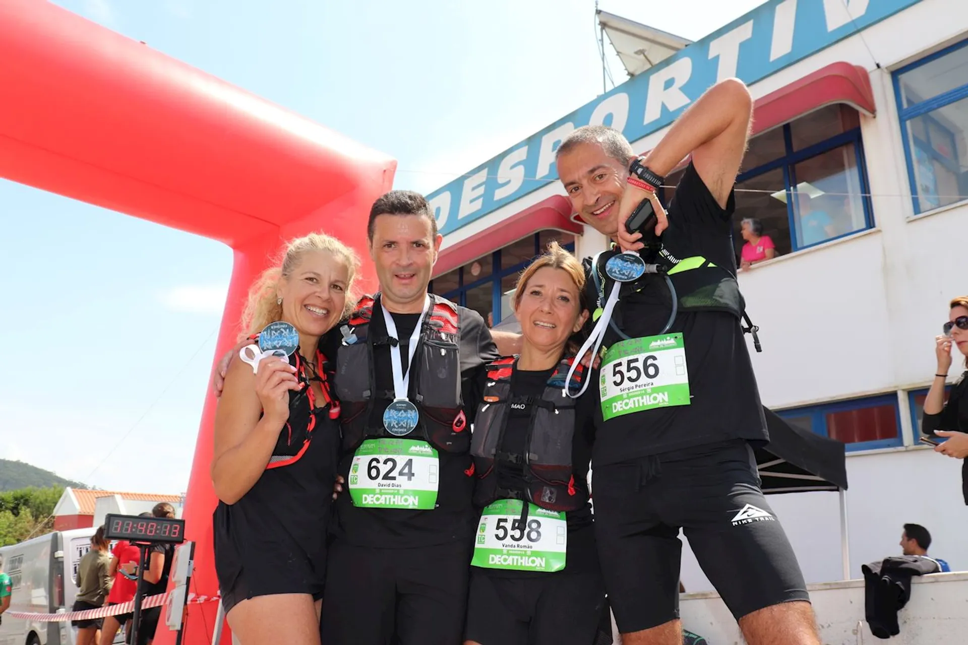 The image shows four runners posing together, each wearing a race bib. They are standing under an inflatable arch, possibly at the finish line of an event. One of the runners is holding up a medal. The background includes a building with the word "SPORTIF" visible. The atmosphere seems celebratory, suggesting they have completed a race.