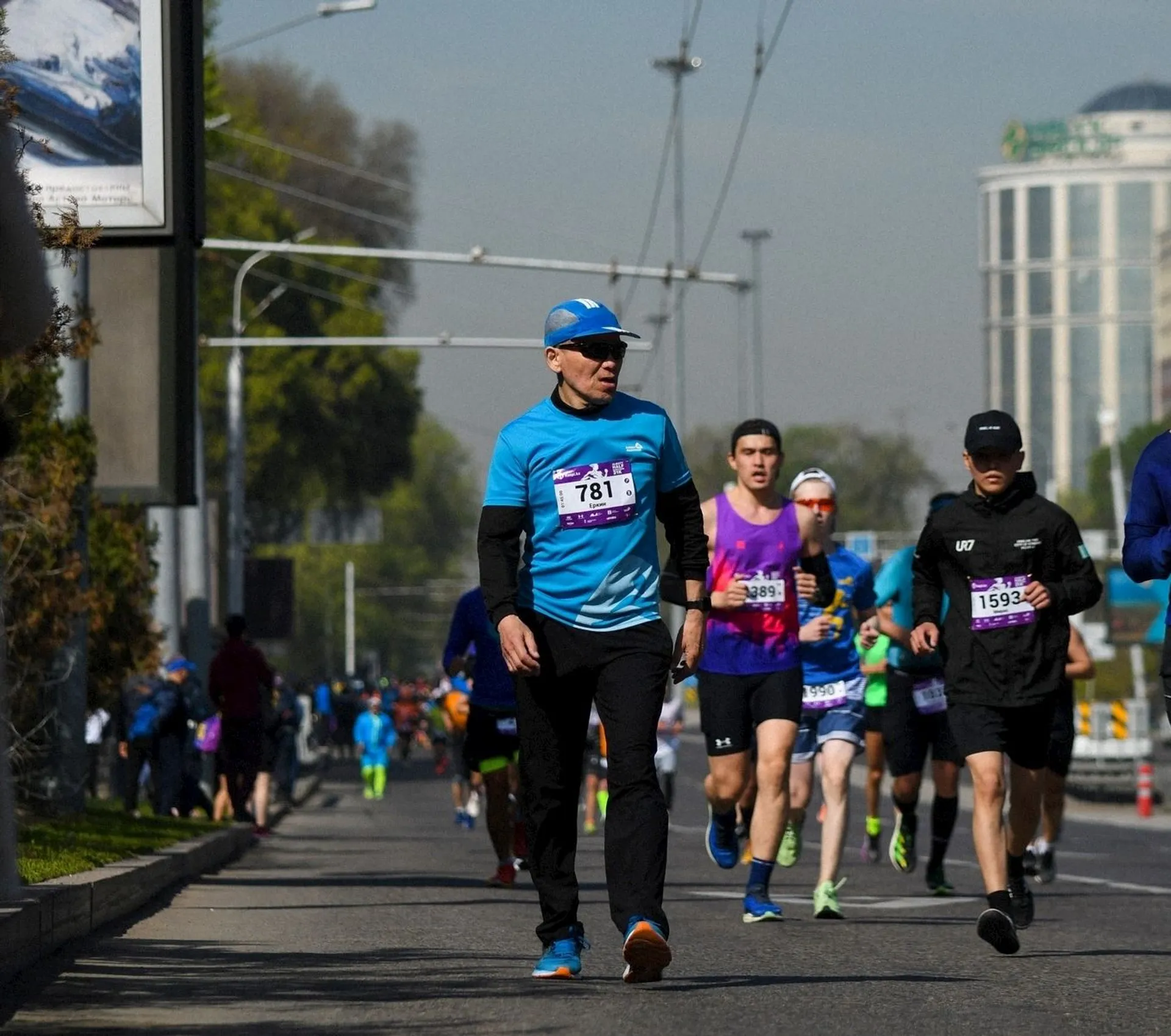 The image shows a group of people running a marathon or a race on a city street. They are wearing athletic clothing and have bib numbers pinned to their shirts. The environment suggests an organized event with several participants.