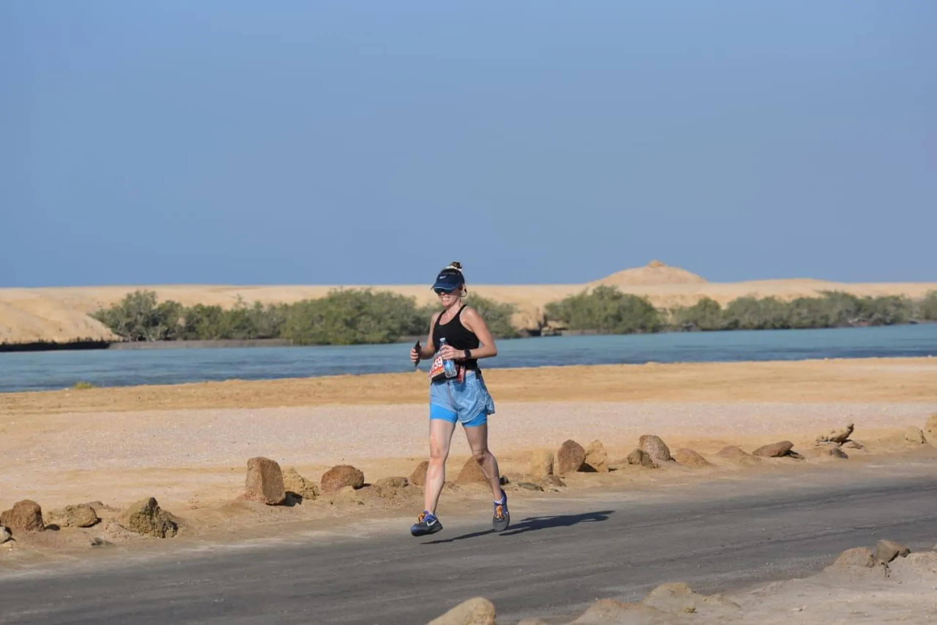 The image shows a person running on a paved road with a bottle in each hand. The background features a body of water and low vegetation. The environment appears to be a desert or arid landscape with clear blue skies.
