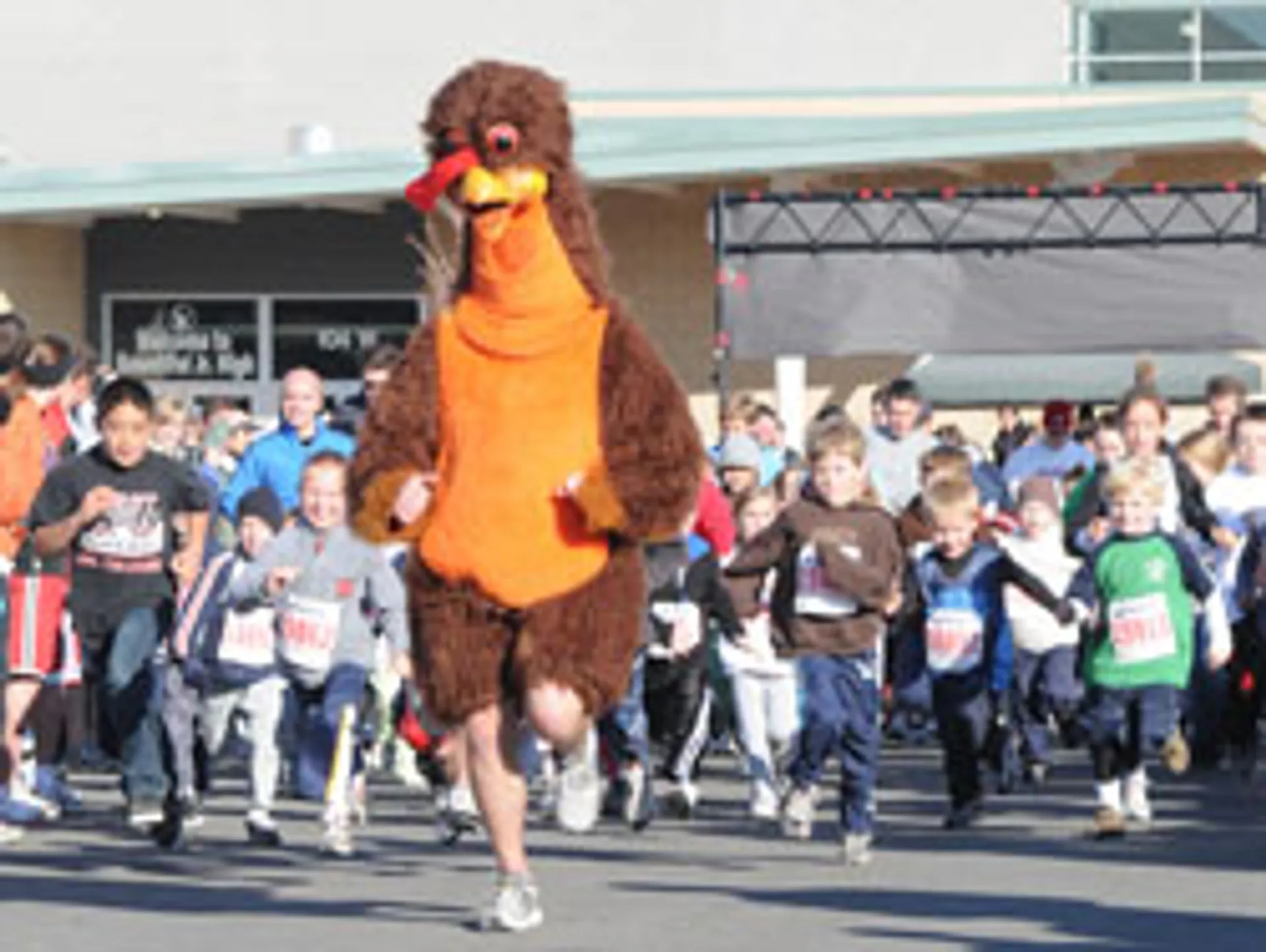 The image shows a person dressed in a turkey costume leading a group of runners at