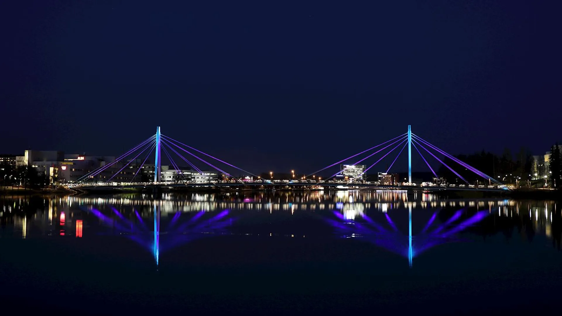 The image shows a nighttime cityscape featuring a calm body of water in the foreground