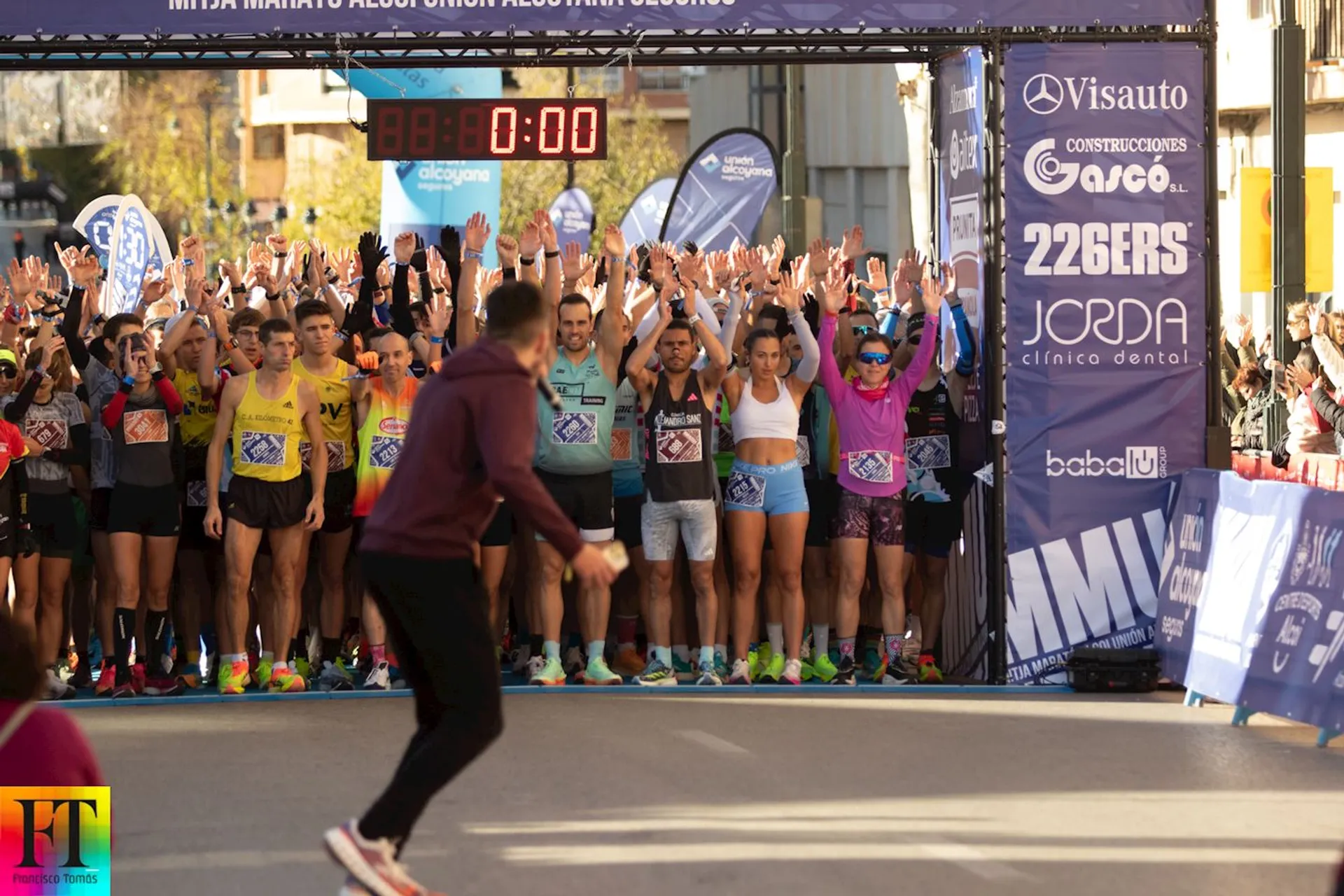 The image shows a group of runners lined up at the starting line of a race. There is a timing clock above the runners showing "00:00", indicating the race is about to start. The participants are raising their hands, and there is a person in the foreground moving away from the camera. Various sponsor banners are visible in the background.