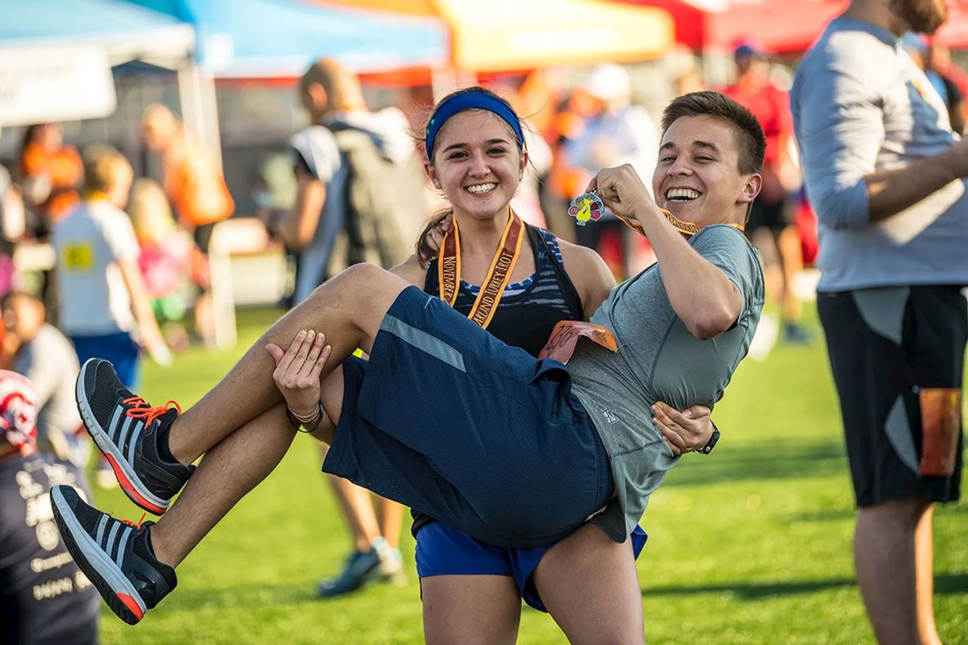 The image shows two people at a sporting event. One person is holding another person, who is displaying a medal. They seem to be celebrating, and there are others in the background also wearing medals. There are colorful tents in the background, likely part of the event setup.