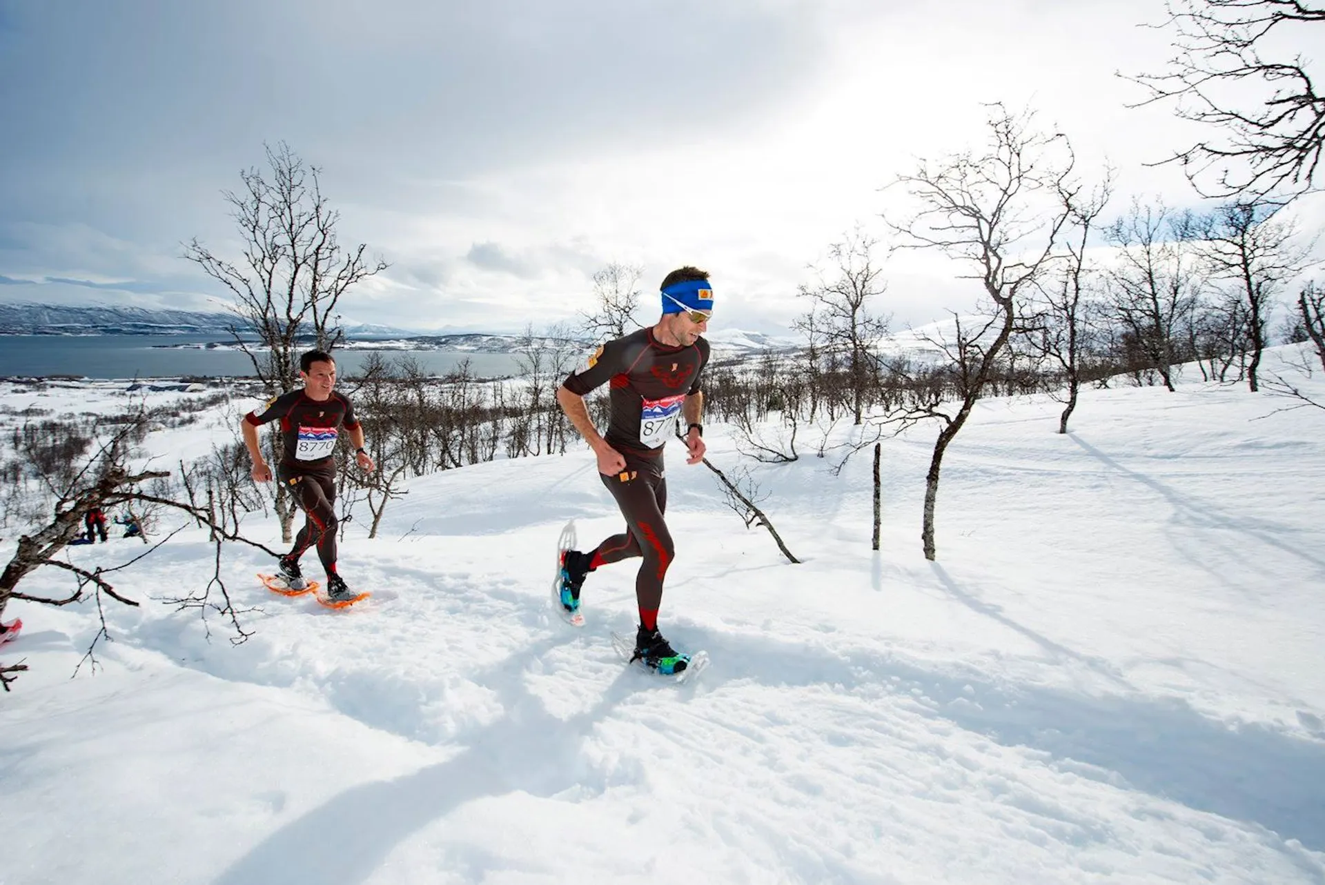The image shows two individuals engaged in a winter running event or race. They are wearing athletic clothing suitable for cold conditions, including long-sleeve tops and running tights or pants. Both runners are equipped with sunglasses and running shoes, and they appear to be running through a snowy landscape with patches of bare trees. The sky is partly cloudy, and a body of water and distant mountains are visible in the background, contributing to a scenic and possibly brisk environment for the event.
