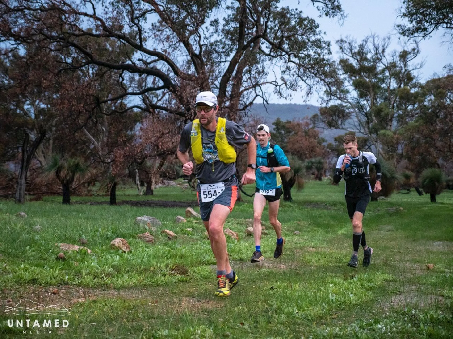 The image shows three individuals participating in a trail running event or race. They appear