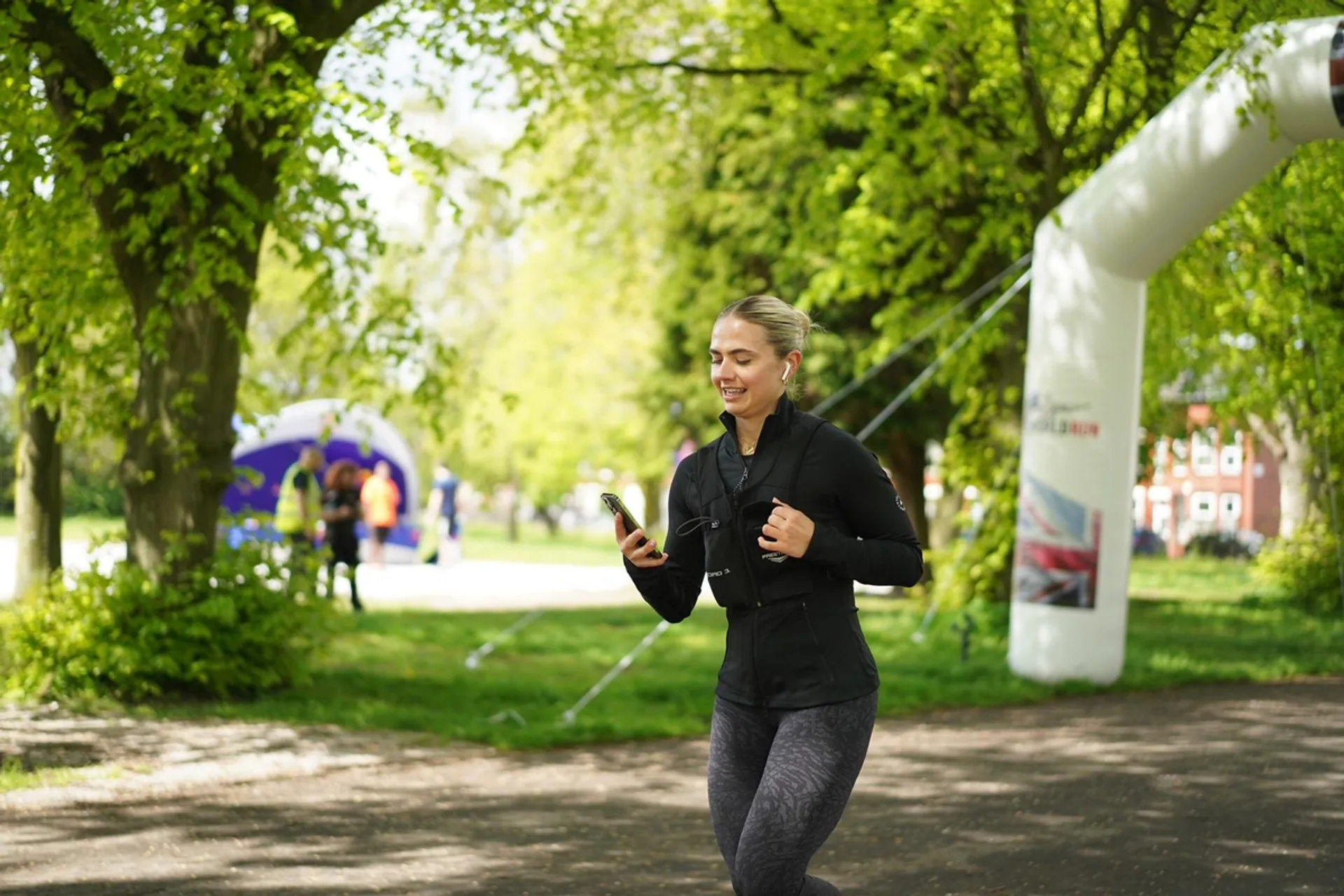 The image shows a woman running outdoors in a park-like setting. She is smiling and appears to be listening to music or using a smartphone, which she is holding in her right hand. She is wearing athletic clothing suitable for running: a long-sleeve top, leggings, and sneakers. There is also a backpack on her back. In the background, there are trees, a few people, and an inflatable arch, which could indicate the presence of a running event or race. The setting looks green and lush, suggesting it might be spring or summer.