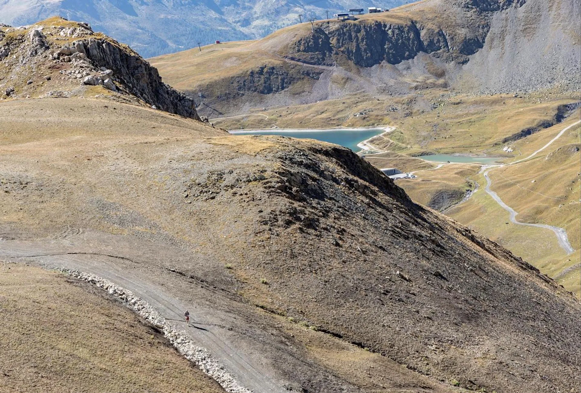 The image shows a mountainous landscape with a distinct ridgeline. In the foreground, there's a narrow path or trail along the ridge with a person walking on it, providing a sense of scale to the vastness of the terrain. In the distance, there are several lakes nestled amidst the mountains, with various shades of blue and green waters. The terrain is rugged and rocky, and it appears to be barren of trees, suggesting a high altitude environment possibly above the tree line. The sky is clear, and the sun is casting sharp shadows, indicating bright and sunny weather conditions. There are also traces of man-made structures, such as roads or paths, in the middle ground, and a building complex at the top right, which could be a