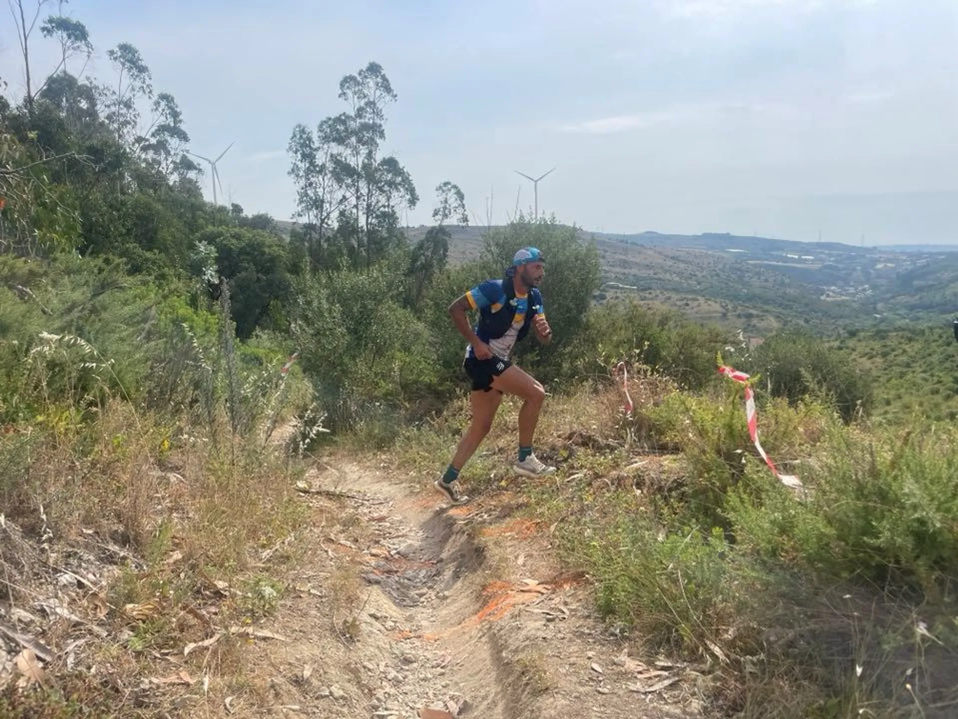 The image shows a person running on a trail in a hilly or mountainous area. The landscape includes vegetation and distant views of hills or valleys. There are also wind turbines visible in the background. The runner is wearing athletic clothing and appears to be participating in a trail running event, as indicated by the markers on the path.