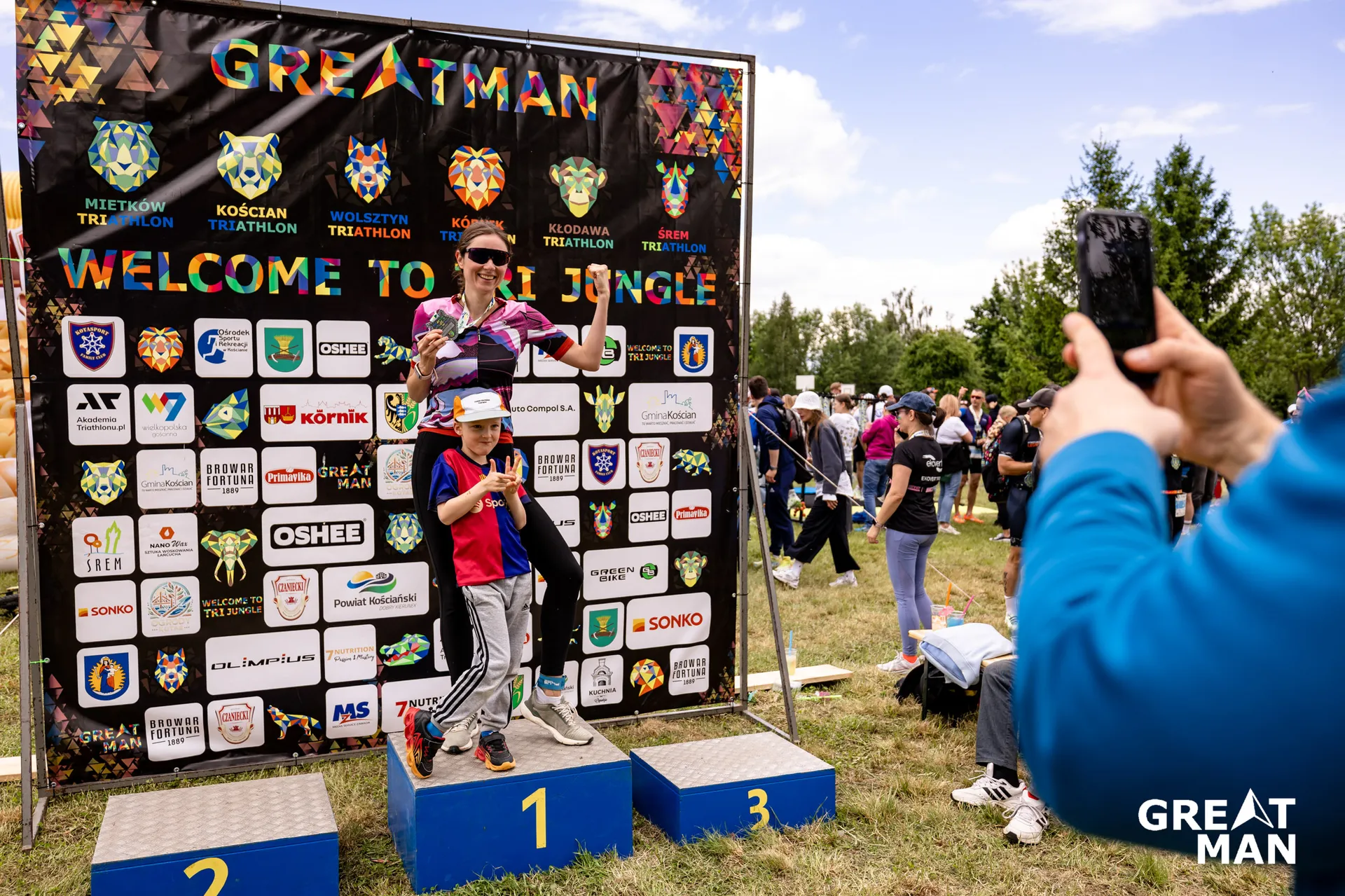 The image shows two people on a podium at the Greatman event. They are posing for a photo in front of a backdrop with event and sponsor logos. The podium is labeled with numbers 1, 2, and 3. There are other people in the background, some taking photos. The scene suggests a celebratory moment at a competition or event.