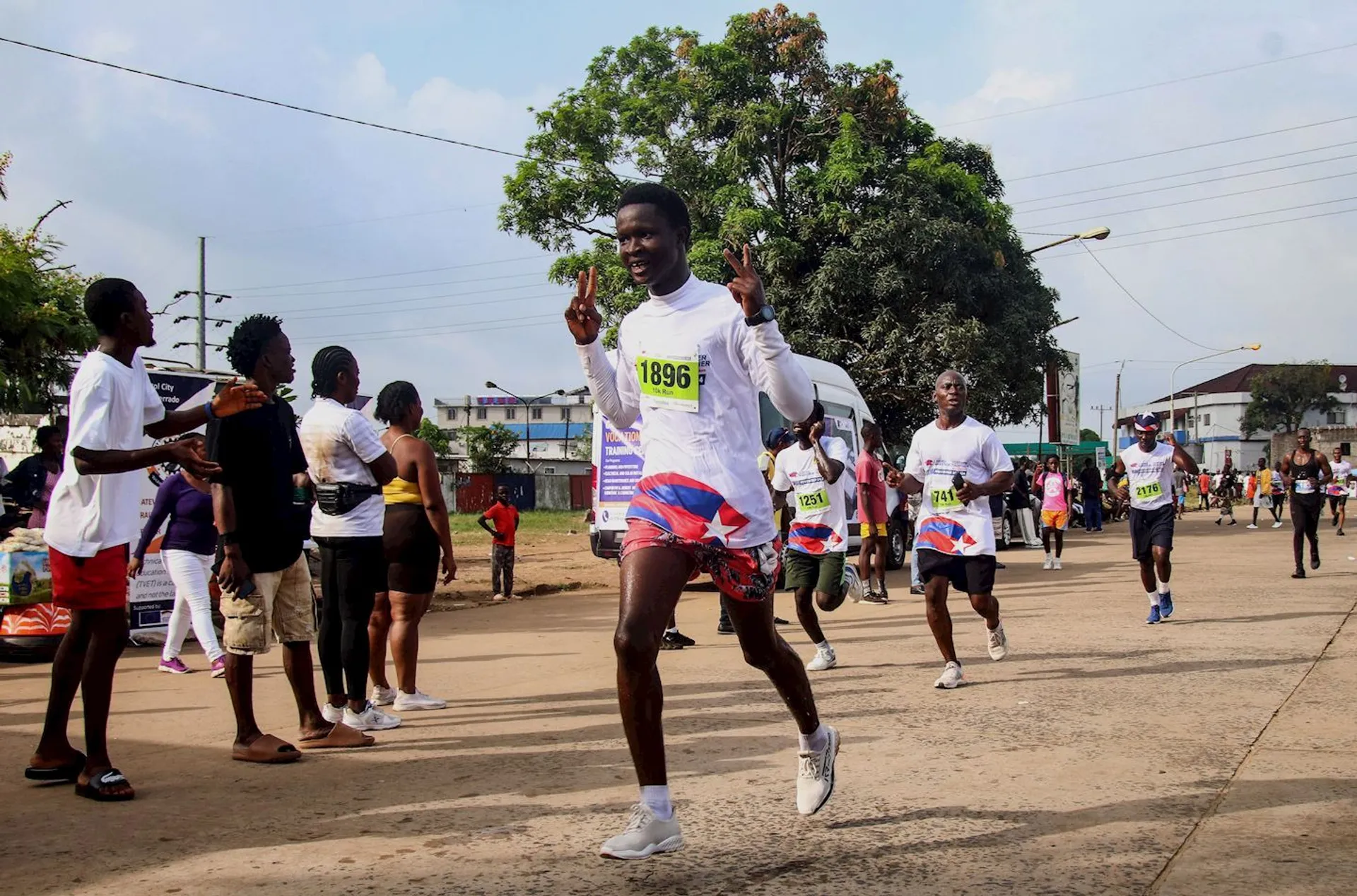 The image shows a group of people participating in a road race or marathon. Runners wearing numbered bibs are on a street, and spectators are watching and cheering them on. The atmosphere is lively, and everyone appears to be enjoying the event.