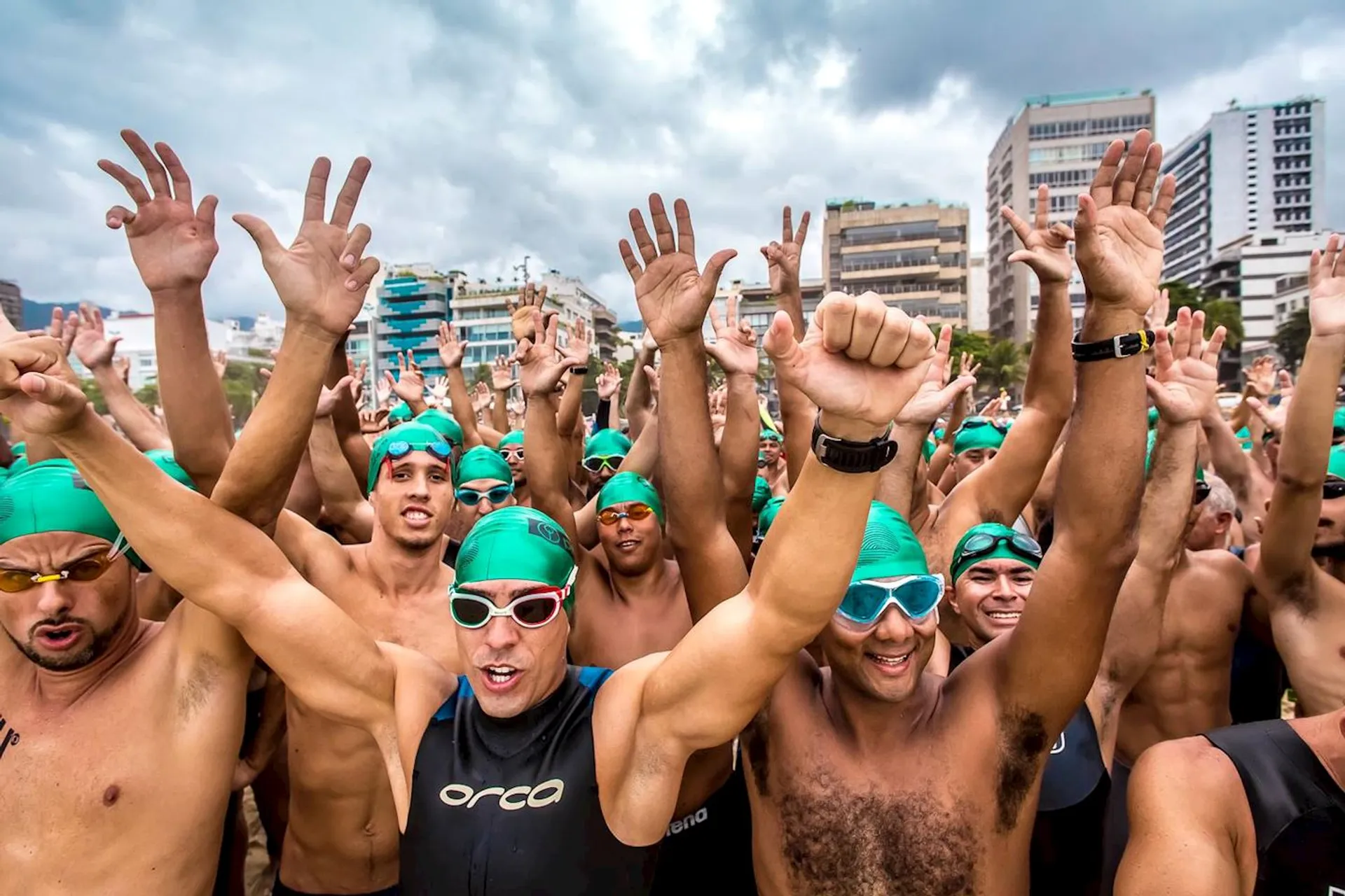 The image shows a large group of people who appear to be preparing for a swimming event. They are wearing swim caps and goggles, and many are raising their hands enthusiastically. The background features a cityscape with various buildings.