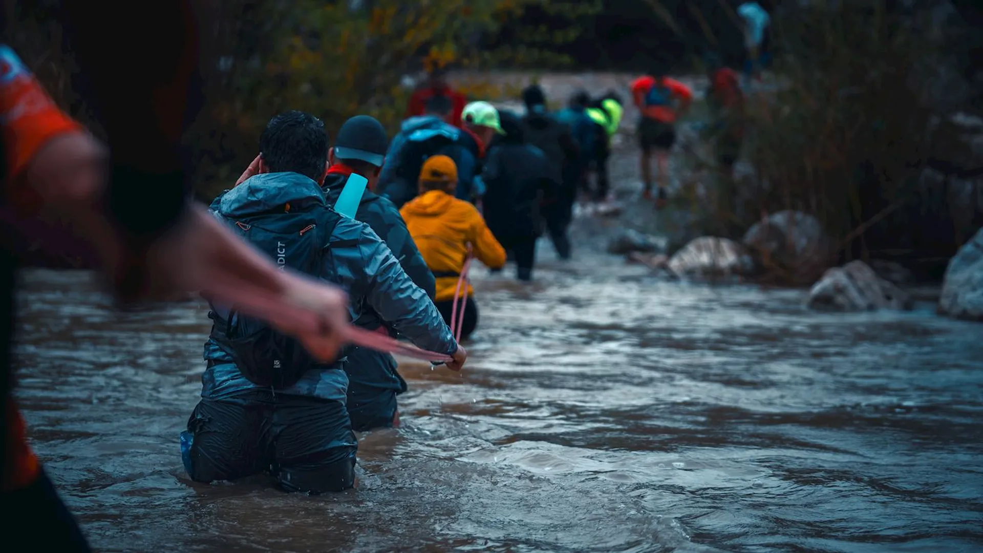 The image shows a group of people wading through a river or stream. They are holding onto a rope for support while moving through the water. The environment appears to be outdoors, possibly during a hiking or adventure activity, with the water level reaching up to their waists in some cases. They are dressed in outdoor or waterproof clothing.