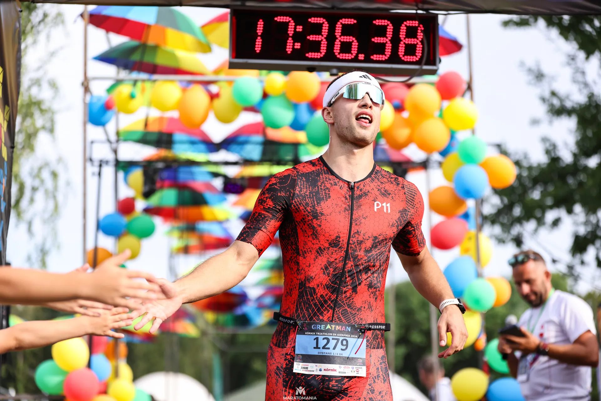 The image shows a person dressed in athletic gear crossing a finish line at an outdoor event. There are colorful balloons and a digital timer displaying "17:36:36" overhead. People are nearby, and some appear to be reaching out to congratulate the participant.