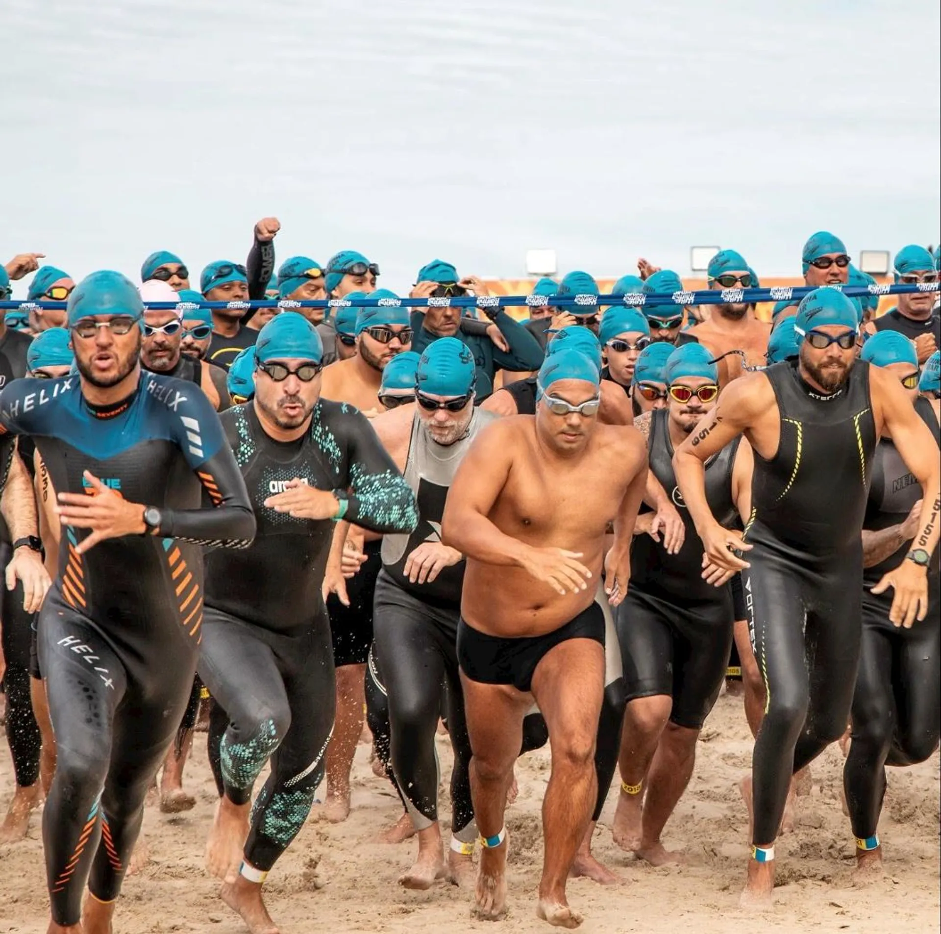 The image shows a large group of people wearing swim caps and wetsuits, running together on a sandy beach. They appear to be starting or participating in a swimming race or triathlon event.
