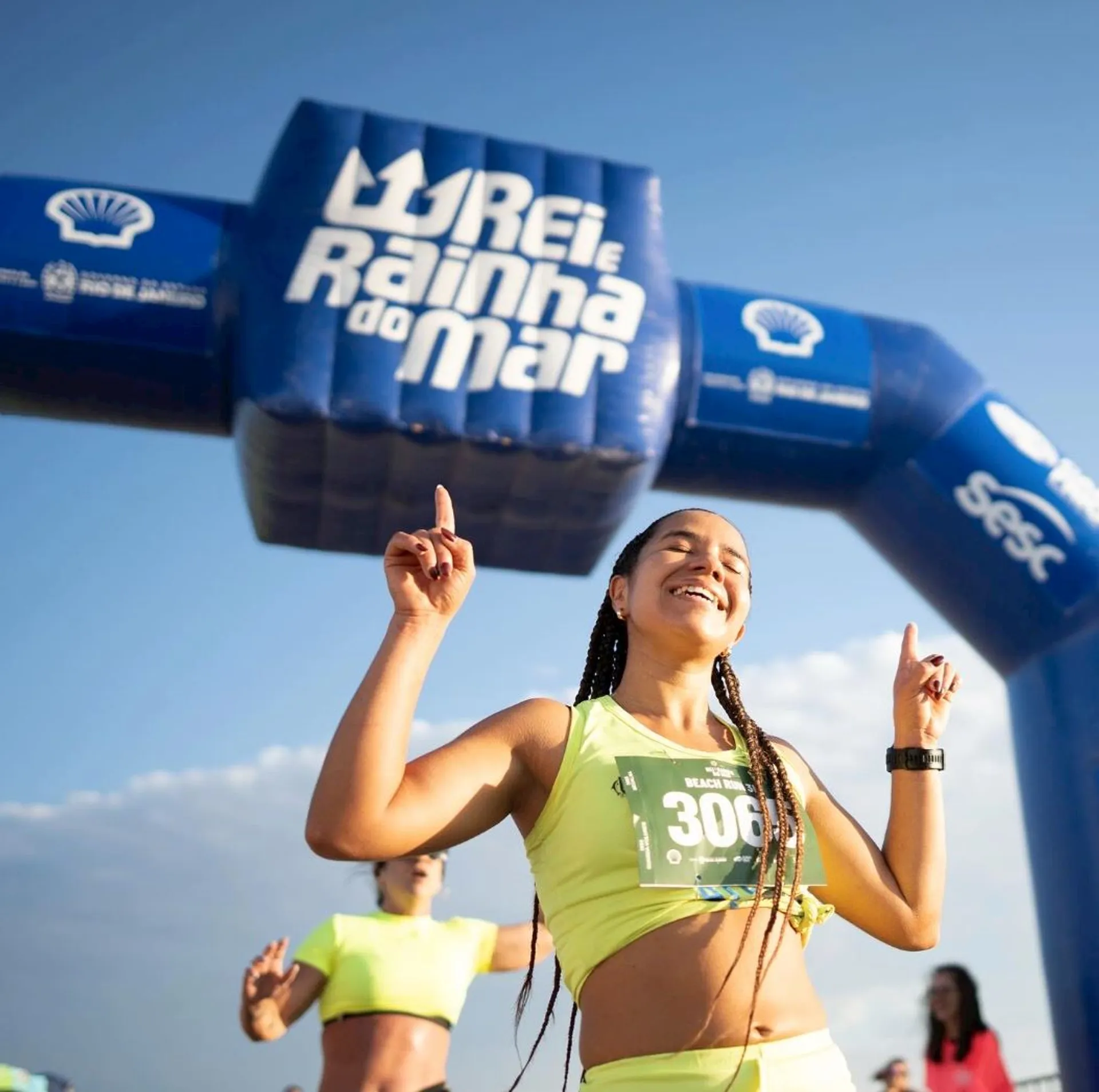 The image shows a woman in athletic clothing smiling and pointing upwards with both hands. She is standing in front of an inflatable arch with the text "Rei e Rainha do Mar," which suggests it's related to a sports event, likely a race or competition.