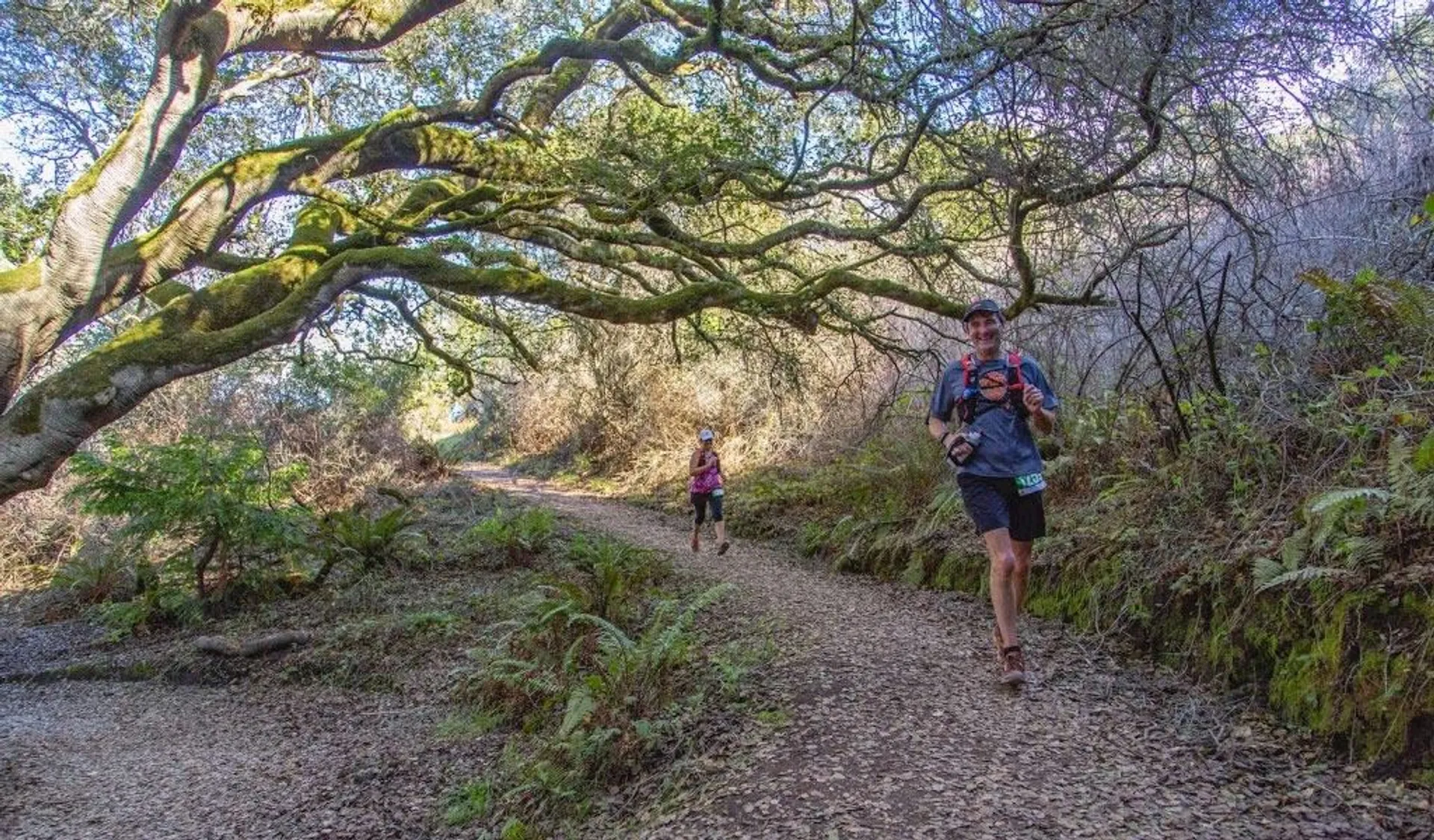 The image depicts two runners on a trail in a natural setting. The foreground shows