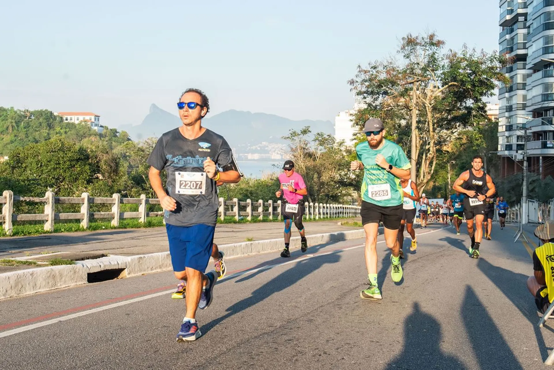 In this image, there are several people participating in a road race or marathon. They are dressed in athletic clothing, and each runner appears to have a bib number attached to their front, which is typical of organized running events. The setting appears to be a sunny day, and they are running on a paved road with some greenery alongside and buildings in the background. There is also a view of a mountain in the distance, indicating the event may be in a scenic or coastal area. It looks like a sports event focused on fitness and outdoor activity.