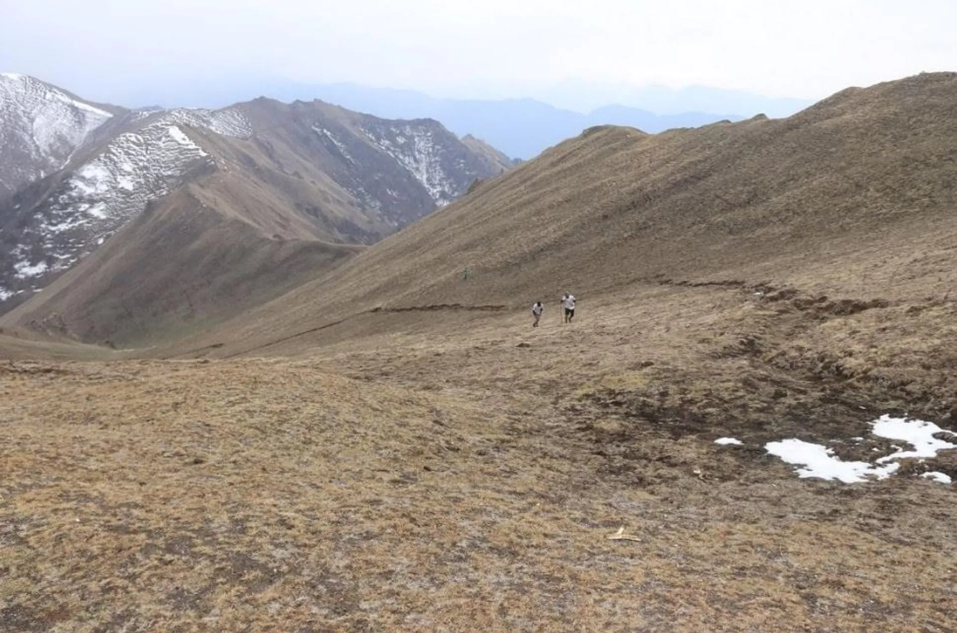 The image shows a mountainous landscape with a few patches of snow. There are three people walking along a path on a grassy and rocky terrain. The mountains in the background have some snow on their peaks, and the scene appears to be taken in a high-altitude area with rugged terrain.