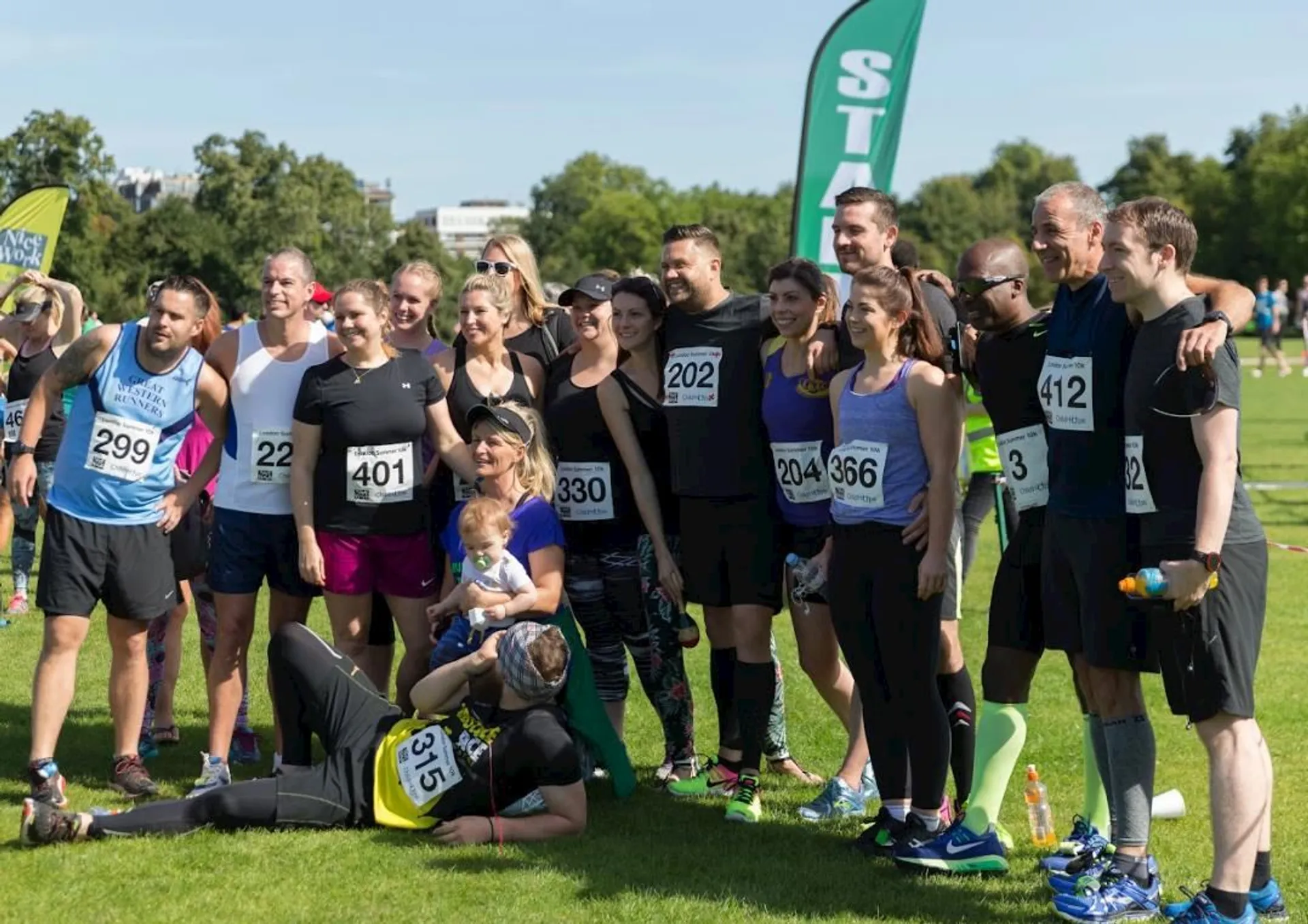 The image shows a group of people gathered outdoors, likely at a running or sporting event, as suggested by the presence of race numbers pinned to some participants' clothes. The participants are a mix of men and women, with varying athletic attire, which implies that they may have just completed a race or a group activity. In the foreground, one person is lying on the grass, possibly stretching or resting, while a child is seen sitting beside an adult, indicating a family-friendly environment. There are banners with the letter "S" visible, probably part of the event branding or sponsorship. It's a bright, sunny day with clear skies, and some trees can be seen in the background, suggesting the event is taking place in a park or open green space