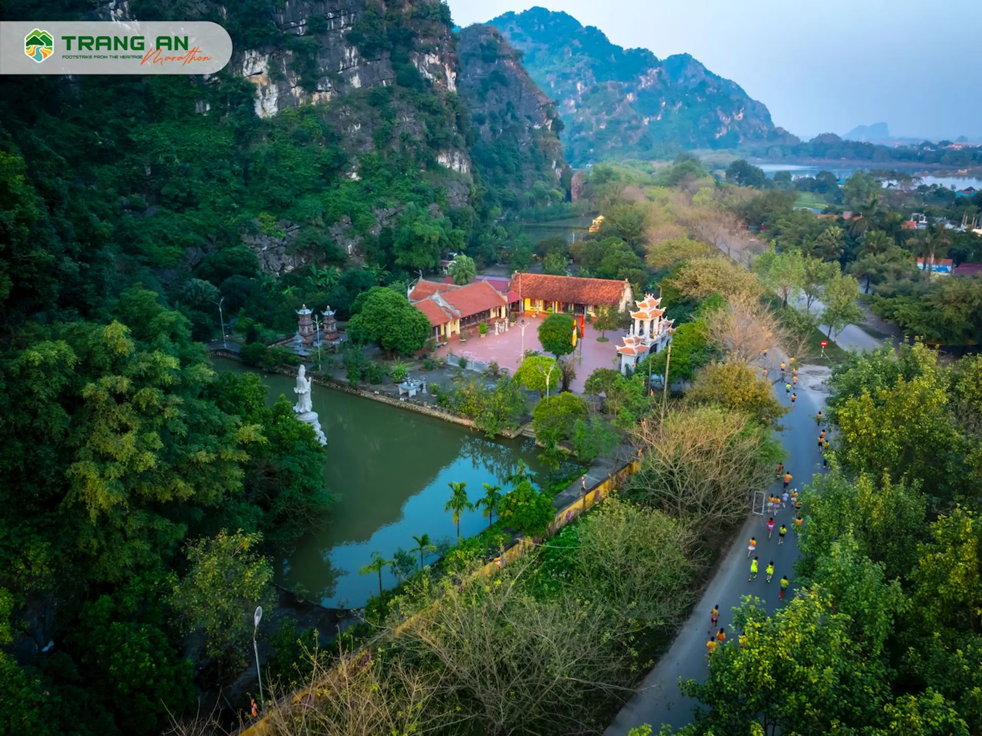 The image shows a scenic aerial view of a lush and verdant landscape. In the center of the image, there's a traditional red-roofed temple or pagoda complex next to a calm body of water that could be a pond or a small lake. There's a reflection of the temple on the water surface, indicating a serene environment. The area is surrounded by rich greenery and towering limestone cliffs or karst mountains, which suggests this location might be in a region known for its natural limestone formations, such as certain areas in Southeast Asia.

There are some people visible on the pathways around the temple and the green area, which might indicate this is a popular spot for visitors or a place of pilgrimage and worship. The road or path winding along