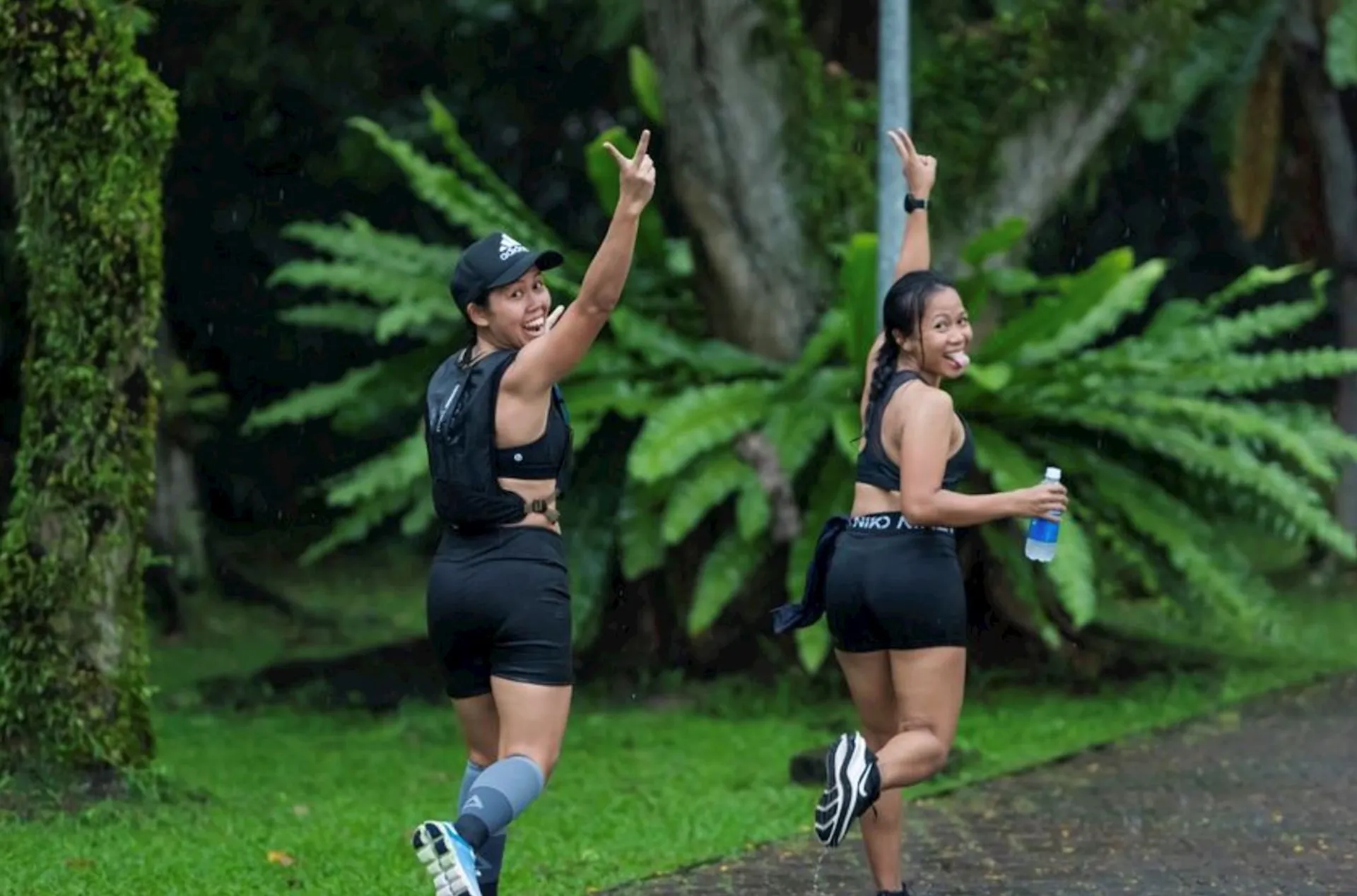 The image shows two individuals who appear to be in a lush, green park or nature area, possibly on a walk or jog. Both of them are wearing exercise attire, with one of them holding a water bottle. They are both smiling and one is making a peace sign with her hand, which suggests they are in good spirits and possibly posing for the photo. The background features large ferns and trees, creating a serene and natural environment.