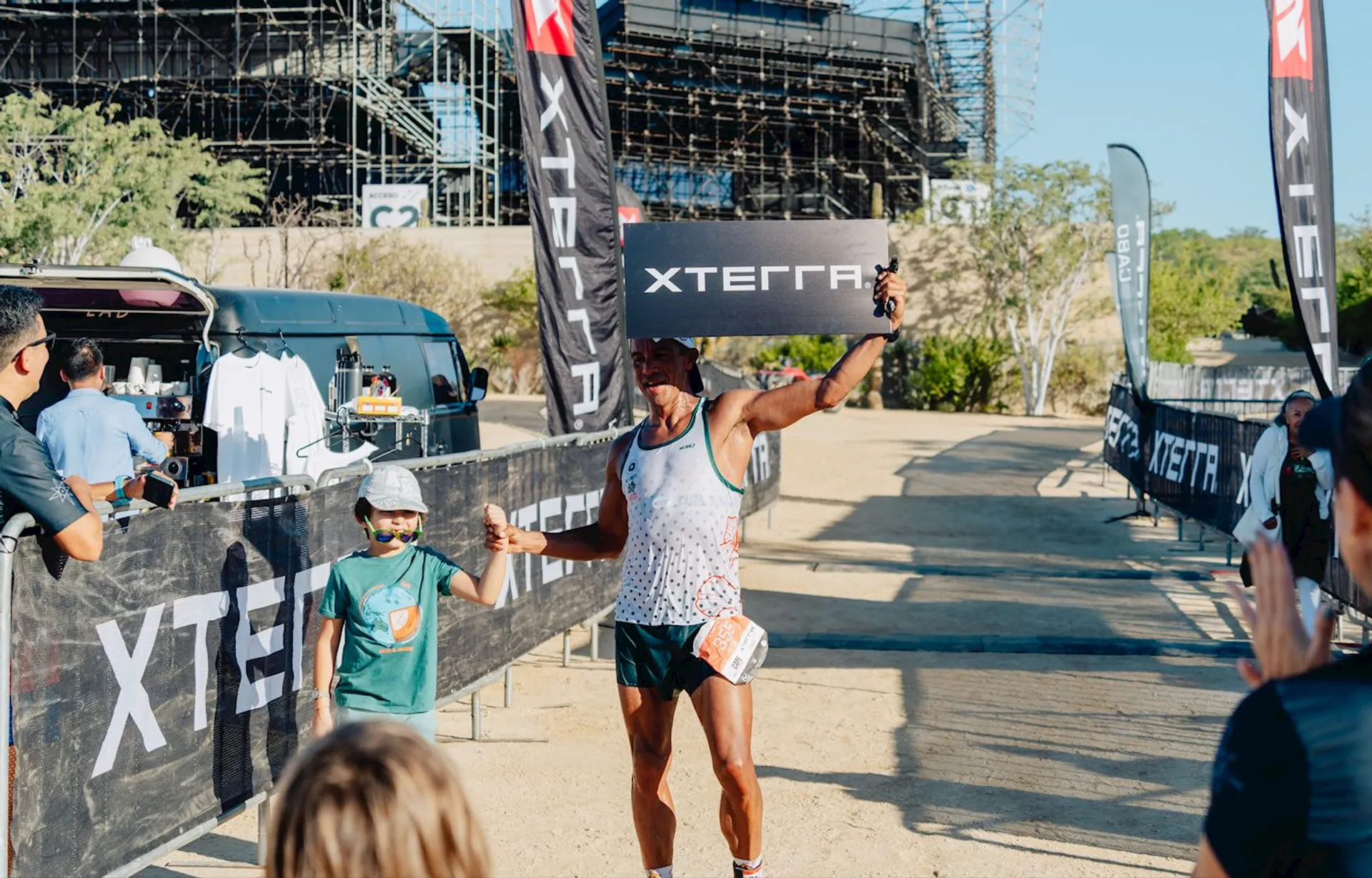 The image shows a person holding an XTERRA sign, finishing a race at an XTERRA event. The scene includes spectators and a young child close by. There are race banners, merchandise, and some scaffolding structures in the background.
