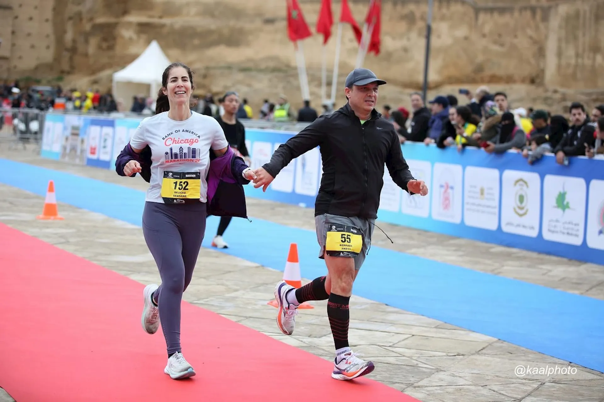 The image shows two people running and holding hands as they cross a race finish line. They are dressed in athletic gear, and there are several banners and flags in the background. It seems to be an organized race event.