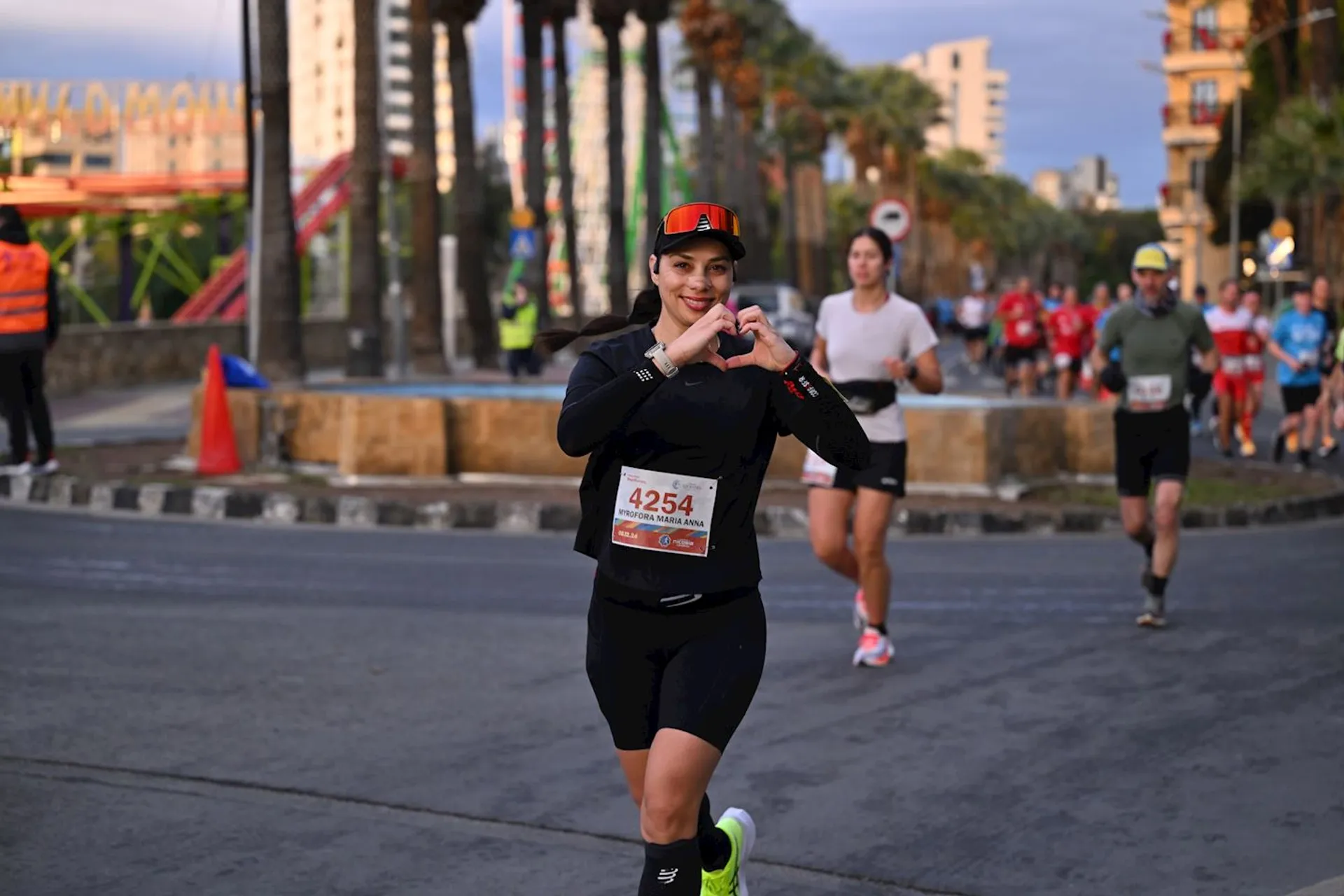The image shows a group of people participating in a running event on a street. The focus is on one individual in the foreground who is making a heart shape with their hands. Other runners and event personnel in safety vests can be seen in the background, along with buildings and palm trees lining the street.