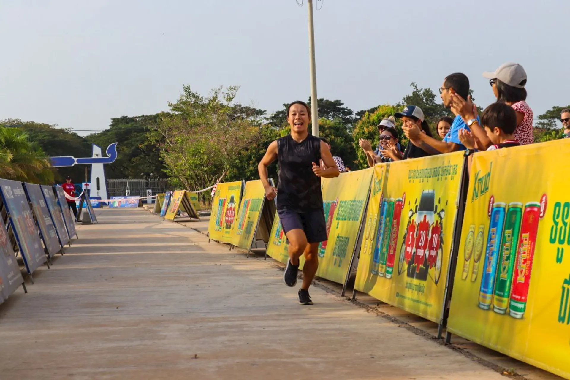 The image shows a person running on a track or path, with spectators on the side cheering them on. There are yellow barriers with advertisements along the path. The setting appears to be outdoors.