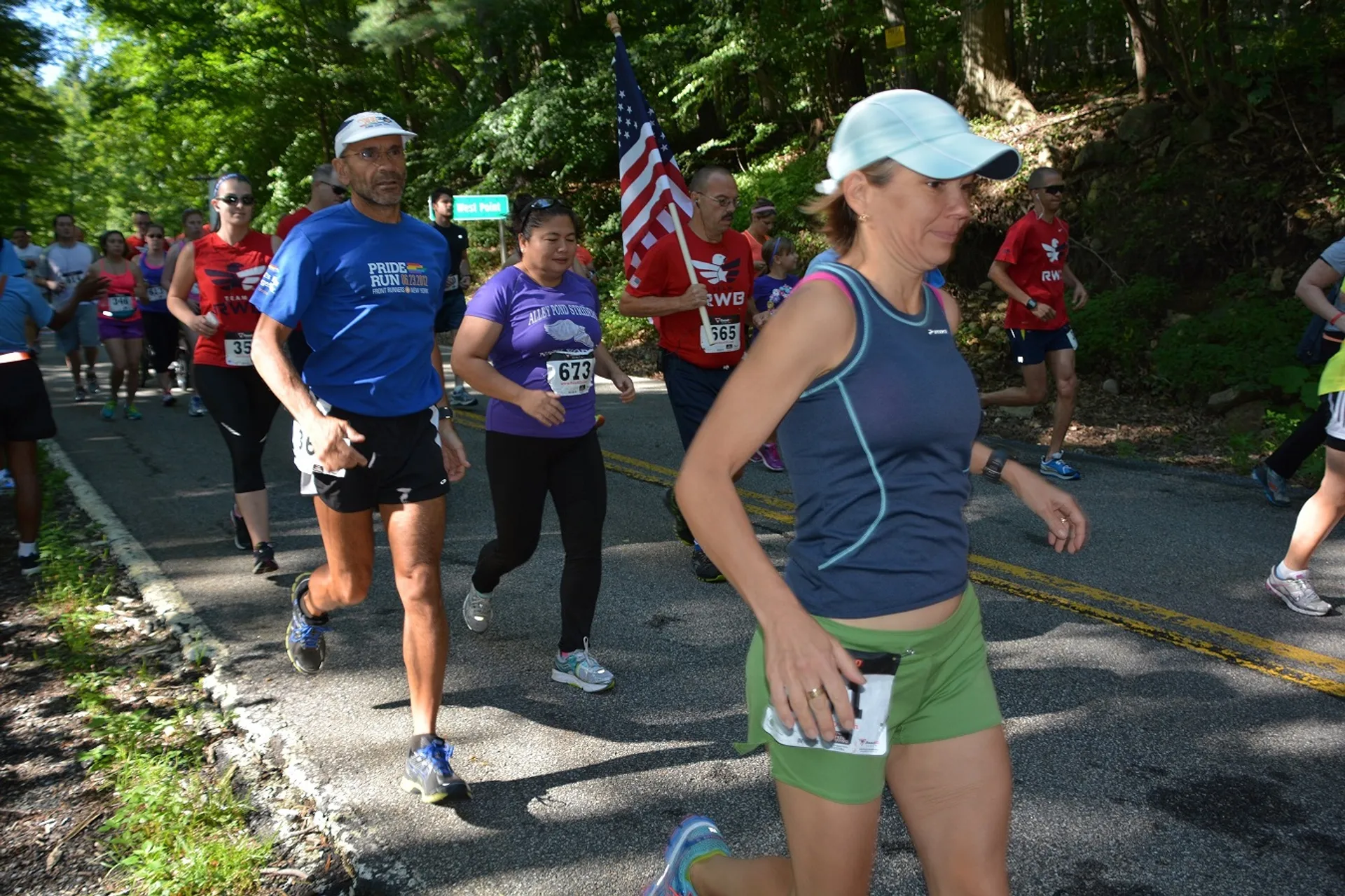The image depicts a group of individuals participating in a running event or race. There are several runners visible, with a variety of running attire indicating a casual, non-professional event. Some runners have bib numbers attached to their shirts, which is typical for organized races. One participant is carrying an American flag, suggesting the event might be associated with a patriotic theme or taking place on a significant national holiday in the United States, such as Memorial Day or the Fourth of July. The environment is verdant and appears to be in a wooded area, indicating the race route may include a scenic or natural path.