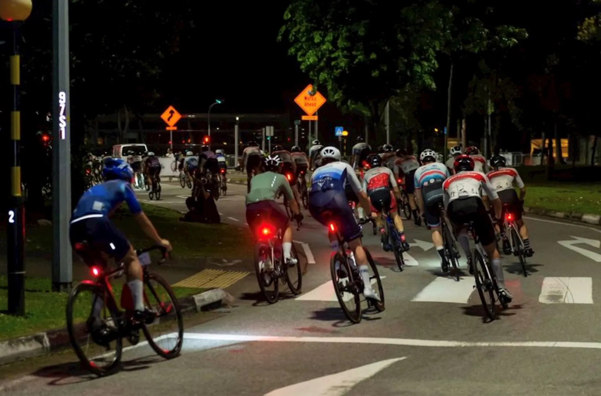 The image shows a group of cyclists riding at night on a street. They are wearing various cycling outfits and helmets, indicating that they might be participating in a group ride or cycling event. The cyclists appear to be riding in a dedicated bike lane or on the right side of the road, following traffic rules. There are street lights illuminating the road, and traffic signs visible, indicating a turn ahead. It seems to be an organized activity given the number of participants and their gear.