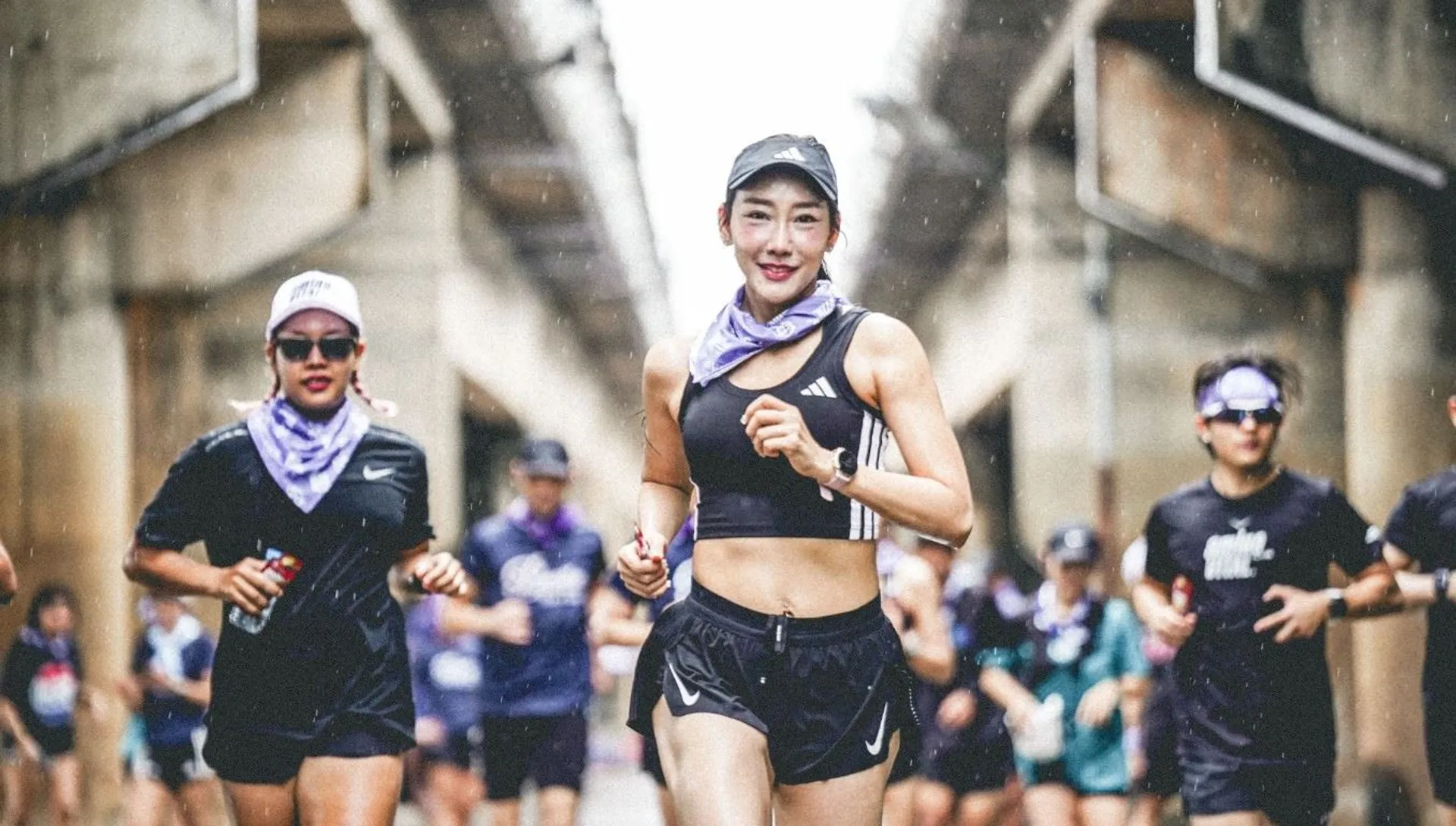 The image shows a group of people running outdoors. The individuals are dressed in athletic gear, and the scene appears to be part of a marathon or group run. The central figure is smiling and wears a cap, a sports bra, a watch, and shorts. There are several other runners in the background.