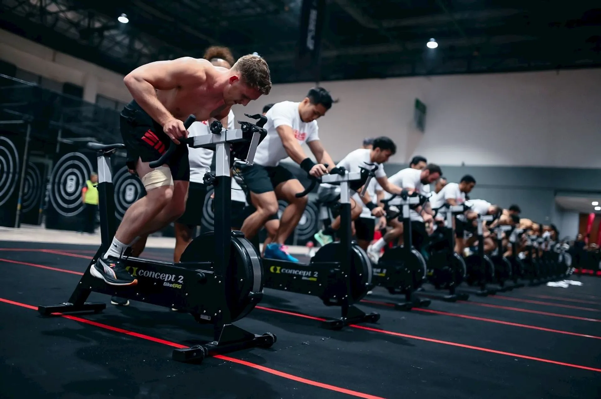 The image shows a group of people working out on stationary bikes in what appears to be a gym or fitness facility. The individuals are aligned in a row, engaged in an intense workout, and the environment suggests a group exercise or spin class.