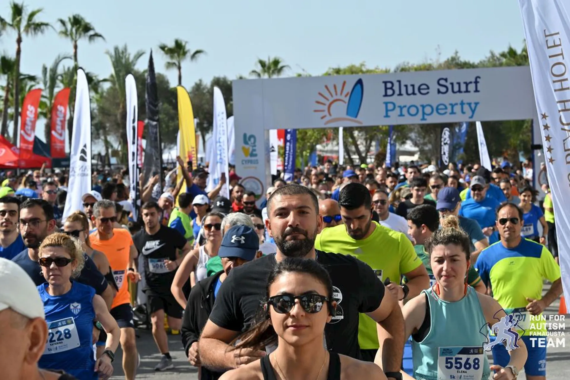 This image shows a large group of people participating in a running event. They are moving under a banner with the text "Blue Surf Property." It's a crowded scene with many runners wearing athletic clothing and numbered bibs. Various banners and flags are visible in the background, indicating different sponsors and organizations.