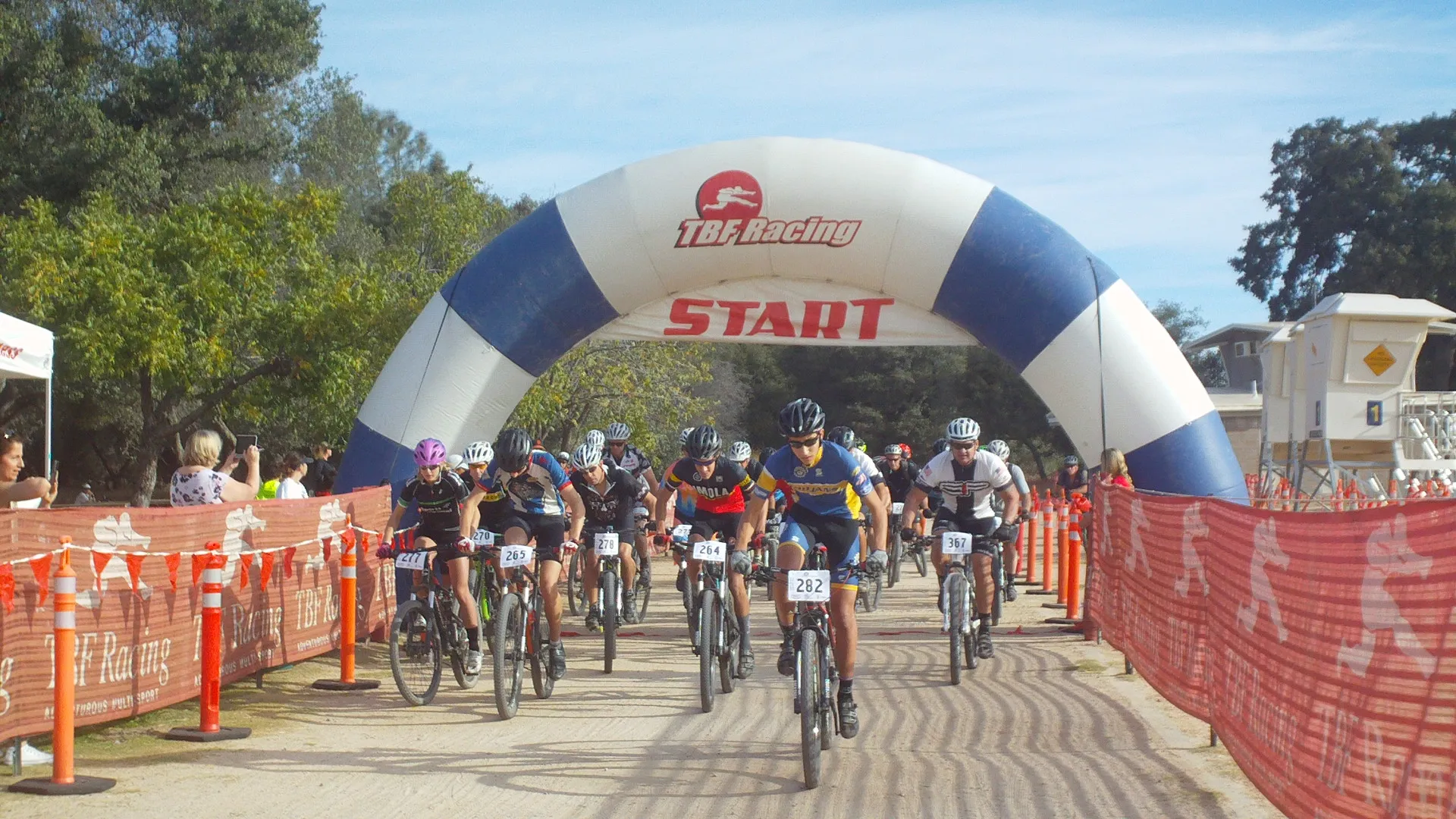 This image depicts a group of mountain bikers at the start of a race. There's a large inflatable arch marked with the word "START" and the logo of TBF Racing, which is likely the organization hosting the event. The bikers are wearing helmets and athletic gear suitable for a race, and are numbered for identification and tracking their race progress. Spectators can be seen on the side of the starting area, behind barriers, likely cheering on the racers. The setting appears to be an outdoor area with trees and grass, indicative of a park or natural trail location commonly used for mountain biking events.