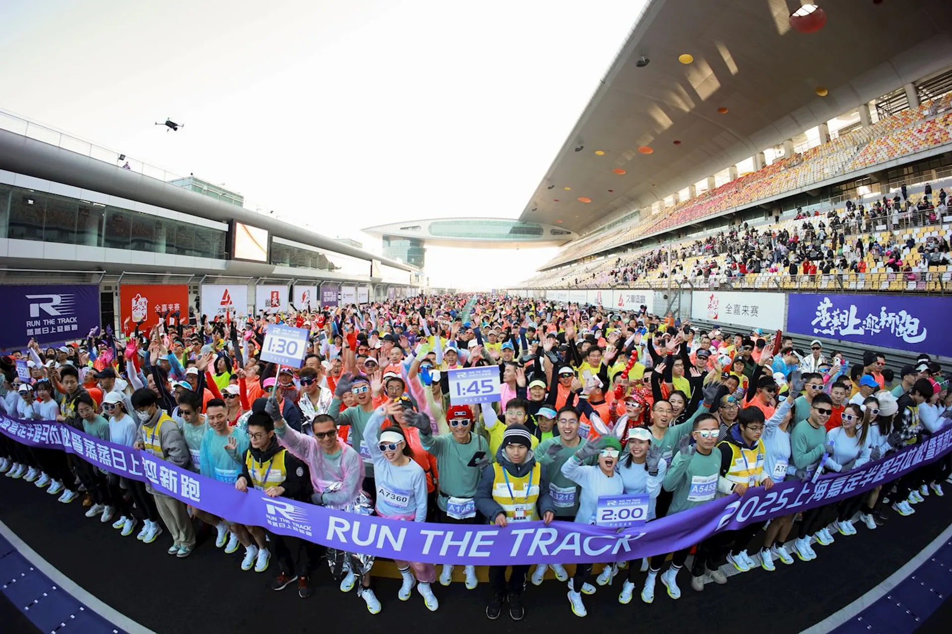 This image shows a large group of people participating in a running event at a race track. Participants are holding signs with expected finish times and wearing bibs, suggesting it's an organized race. They seem to be in high spirits, as many are waving or raising their hands. Banners and advertisements are visible along the sides of the track.