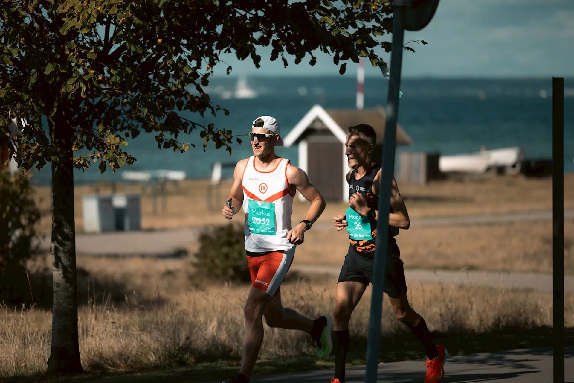 The image shows two people running outdoors near a body of water. They appear to be participating in a race, as they are wearing numbered bibs. The setting is a sunny day with a tree in the foreground and some buildings and boats in the background.