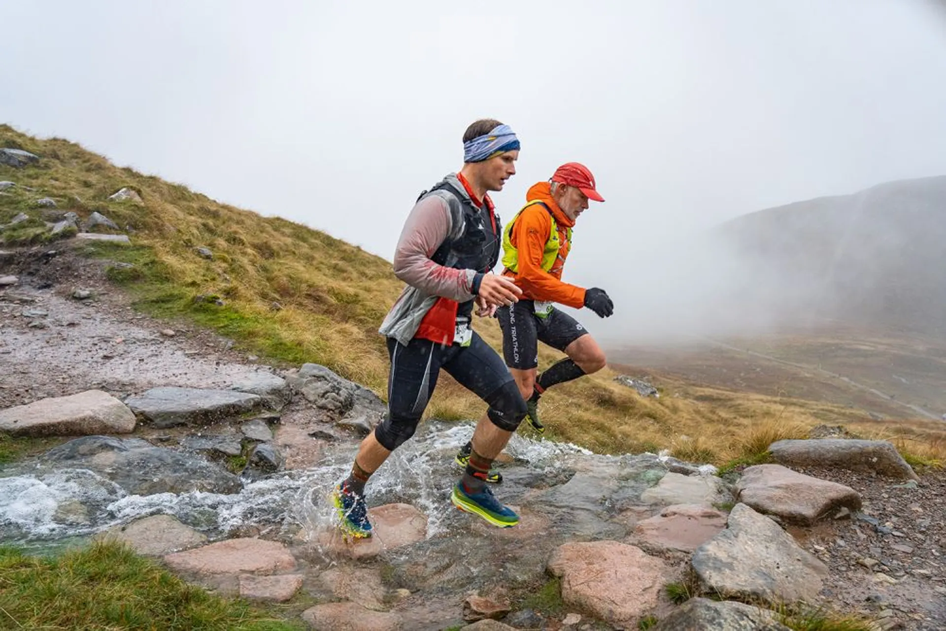 In the image, there are two individuals engaged in trail running. They are mid-stride, crossing a small stream with water splashing around their feet, indicating movement. Both are wearing athletic gear suitable for the activity and the conditions; one person is wearing a blue and gray outfit with a cap, while the other is wearing a bright orange windbreaker and a red hat. The scenery suggests they are in a mountainous or hilly outdoor area with a foggy atmosphere, which could indicate higher altitude or early morning conditions. The terrain appears rocky and uneven, typical for trail running environments.