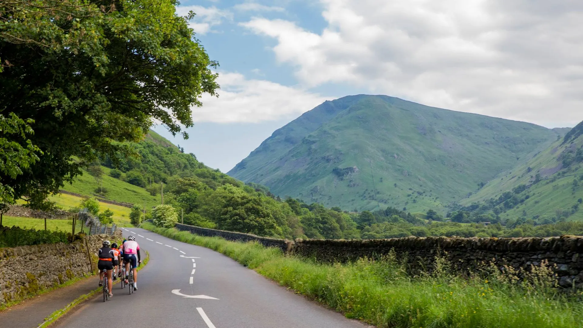 The image shows a group of cyclists riding along a road that cuts through a picturesque rural landscape. On the right side of the road, there's a stone wall typical of the countryside, and beyond it, you can see lush green fields and scattered trees. Dominating the background is a large hill or mountain with a smooth, grass-covered slope, characteristic of a region with rolling hills or a mountainous area. The weather appears to be clear and sunny with a few clouds in the sky, which suggests a pleasant day for outdoor activities like cycling.