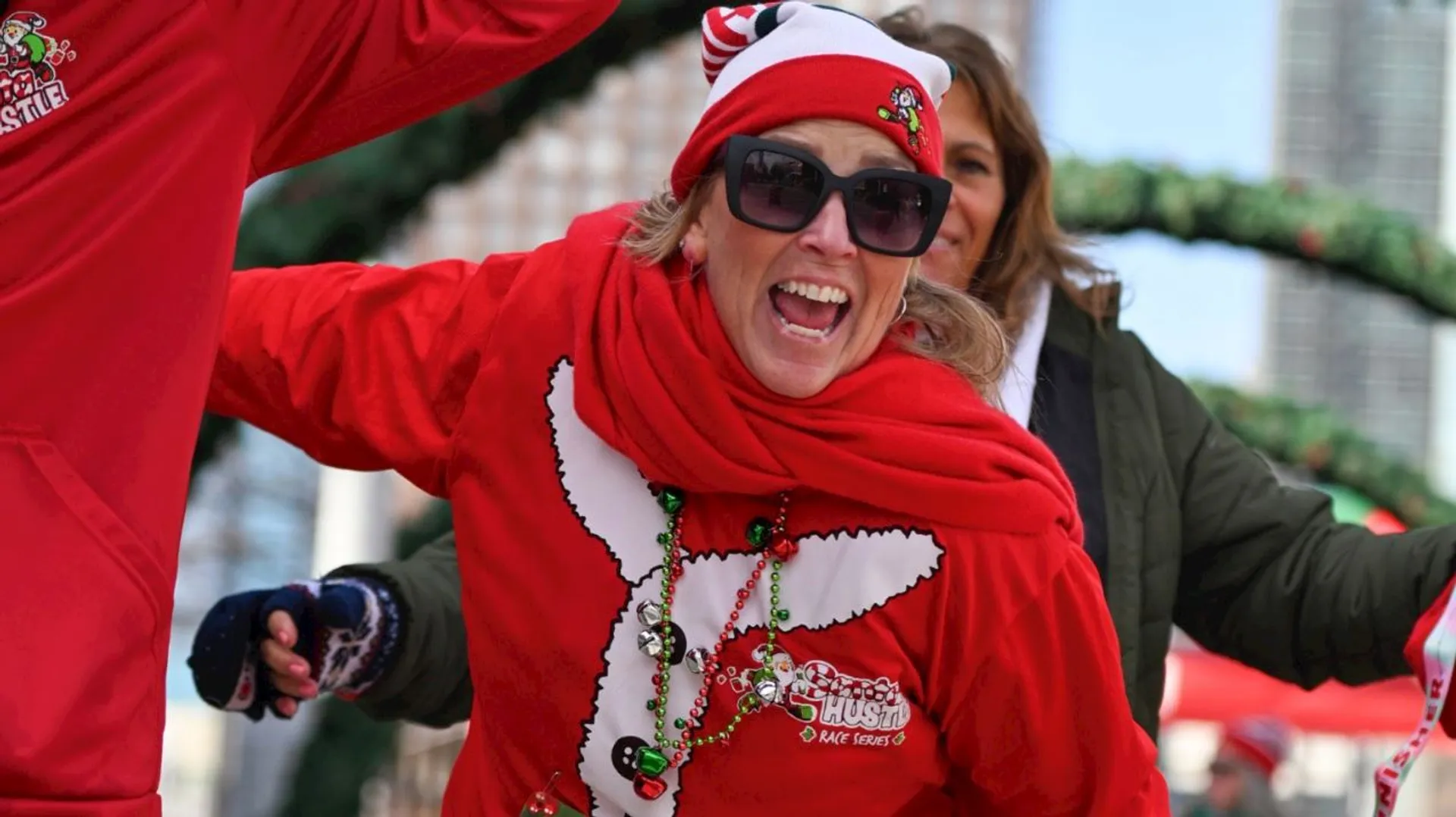 The image shows a person dressed in festive holiday attire, wearing a red sweater, hat, and scarf, along with sunglasses. There are holiday-themed decorations on the outfit and people in the background wearing similar attire, suggesting a festive event or parade.