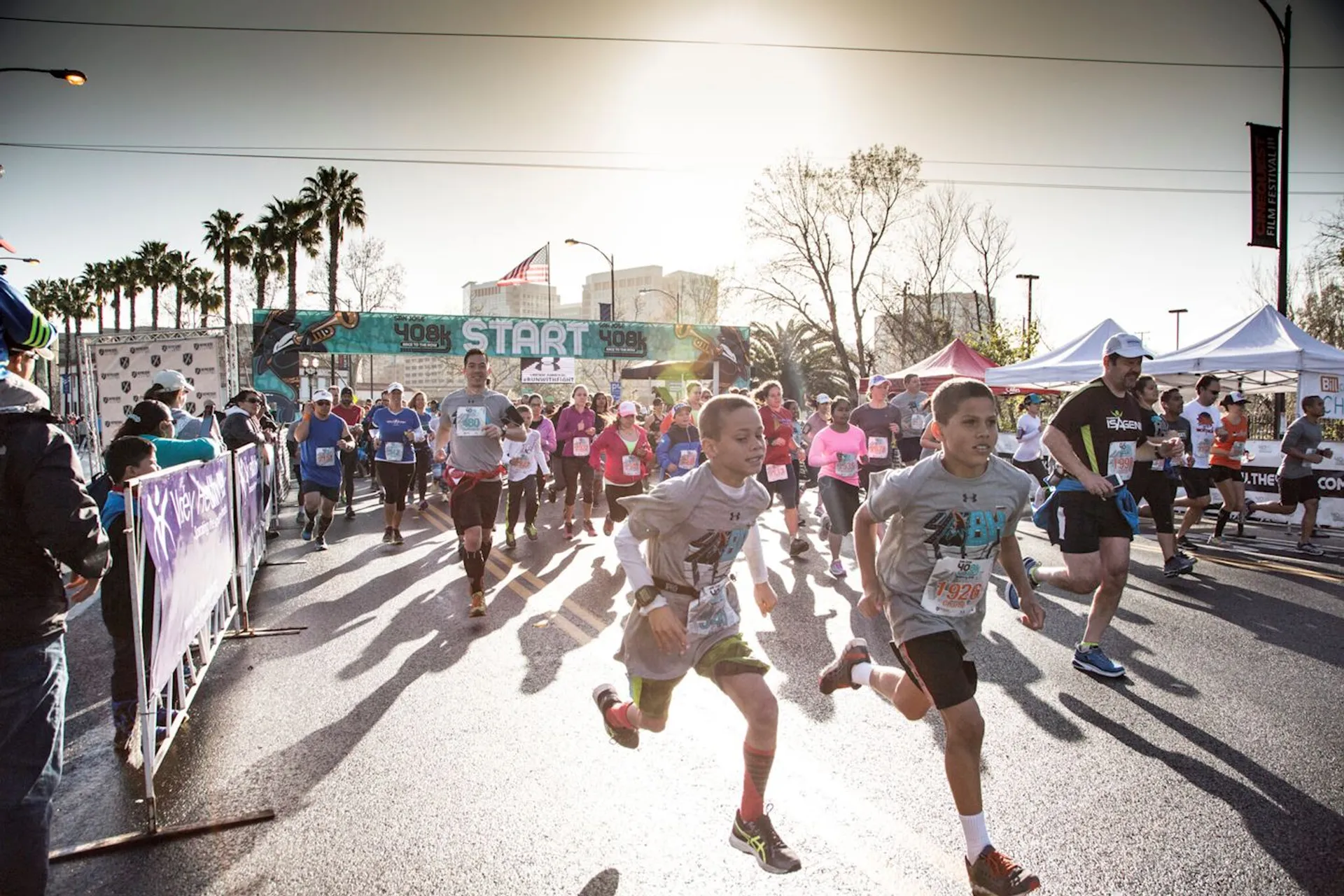The image shows a group of people participating in a running event. They're near a starting line, marked by a "START" banner. The scene is vibrant, with participants of various ages wearing race bibs. There are tents and spectators in the background, and the sun is shining brightly, casting shadows on the road.