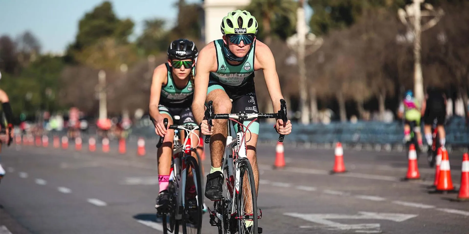 The image shows two individuals on racing bicycles, dressed in typical cycling or triathlon gear, which includes helmets, sunglasses, and form-fitting athletic clothing suitable for cycling and potentially for swimming and running if they are participating in a triathlon. They appear to be part of a competitive event, as suggested by the race bibs on their clothing and the numbered cones lining the route. This setting indicates they might be on a closed road specifically designated for a race. The focus and determination on their faces imply that they are competing or training with serious intent. The background is blurred with trees and a clear sky, conveying a sense of motion and speed.