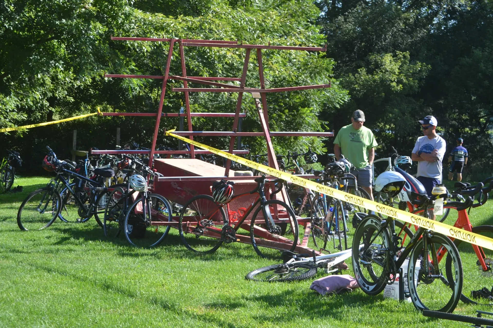 In this image, you can see a variety of bicycles parked on a grassy area, with two individuals standing close by. There is also a large, unique, multi-tiered bike rack structure with several levels for securing bicycles. It seems to be a sunny day and the setting appears to be a park or a similarly green outdoor area. The area is cordoned off with yellow caution tape, suggesting that this may be part of an event or a special area designated for bike parking. The people present are dressed in casual, athletic attire suitable for biking.