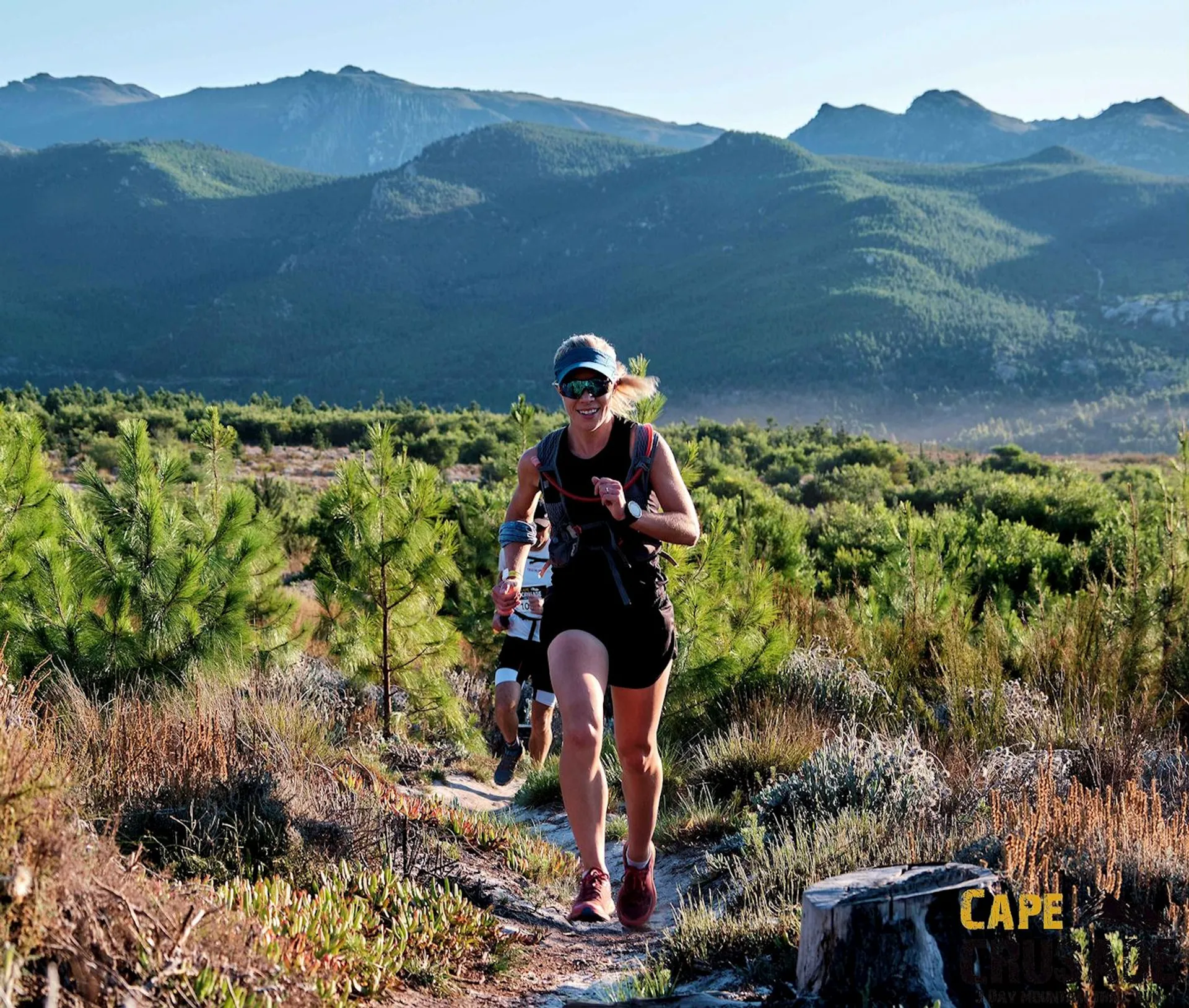 The image depicts a woman trail running in a natural setting. She appears to be