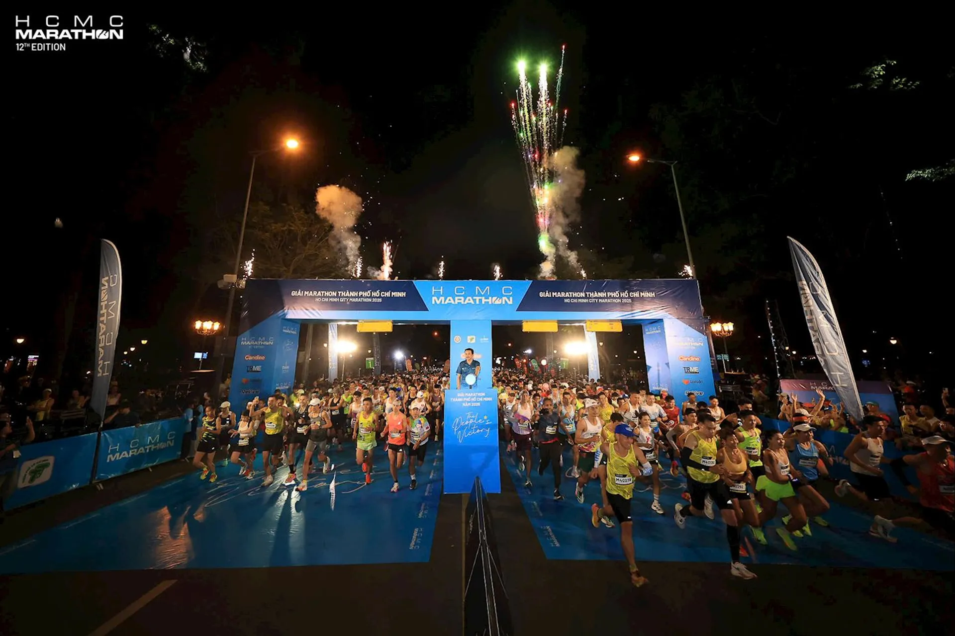 The image shows the start of a marathon event at night. There are many runners beginning the race from a starting line under a blue archway that reads "HCMC Marathon 12th Edition". Fireworks are visible in the sky above, and the area is well-lit with bright lights. Banners and signage related to the marathon are present around the start line.